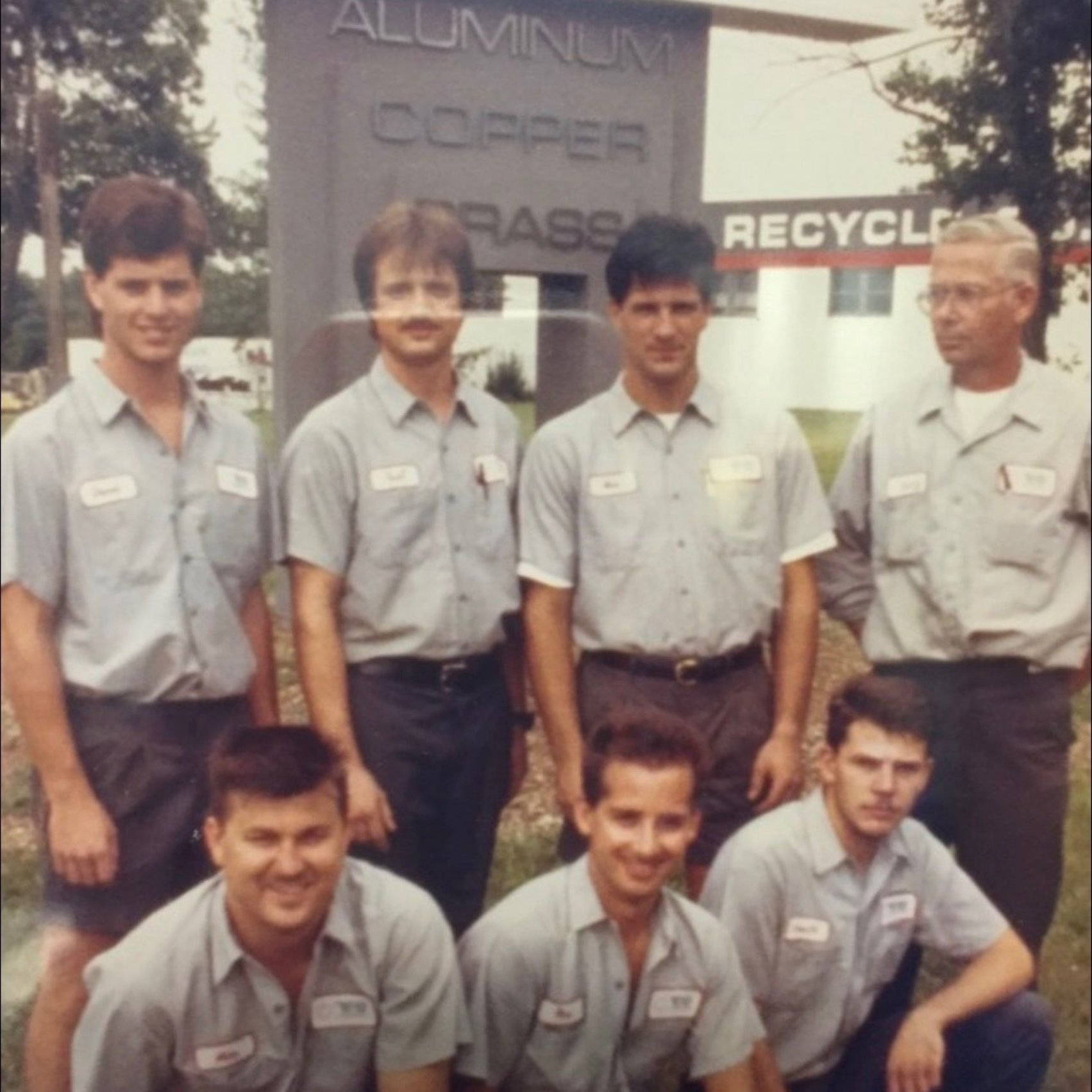 Seven men in matching gray work uniforms pose outdoors in front of a recycling facility sign.