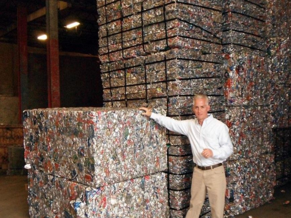A person stands in a warehouse next to large, stacked bales of crushed, colorful recycled aluminum cans.
