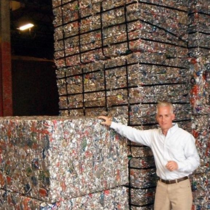 A person stands next to several large, stacked bales of compressed aluminum beverage cans in an industrial warehouse.