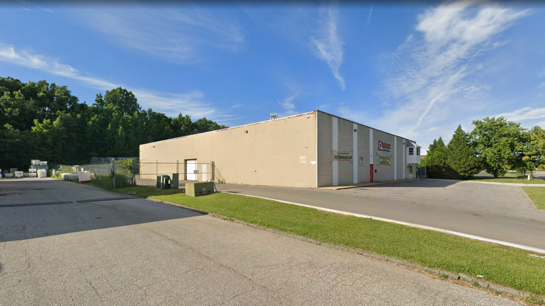 A beige, rectangular industrial or warehouse building under a blue sky, surrounded by asphalt and a grassy area.