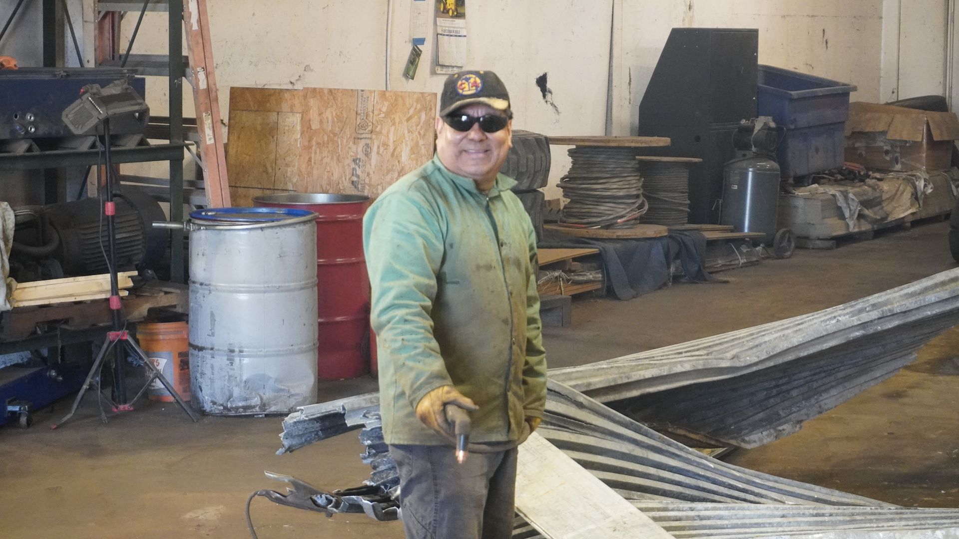 Worker in green jacket and cap standing in an industrial workshop beside stacked metal sheets.