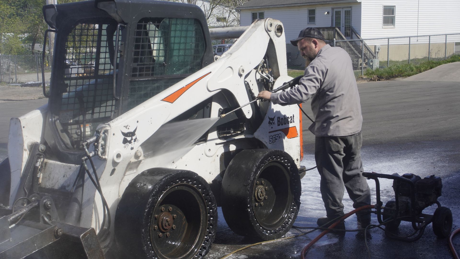 Person pressure-washes a white Bobcat skid steer outdoors on pavement.