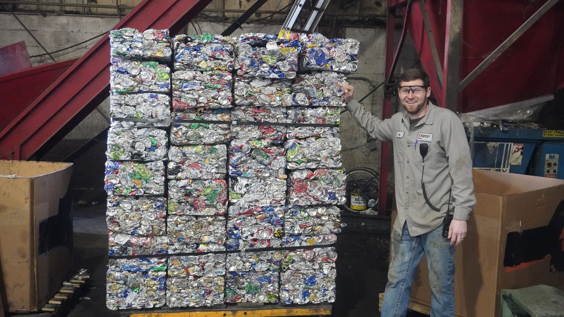 Man standing beside a tall stack of compressed cans in an indoor recycling facility