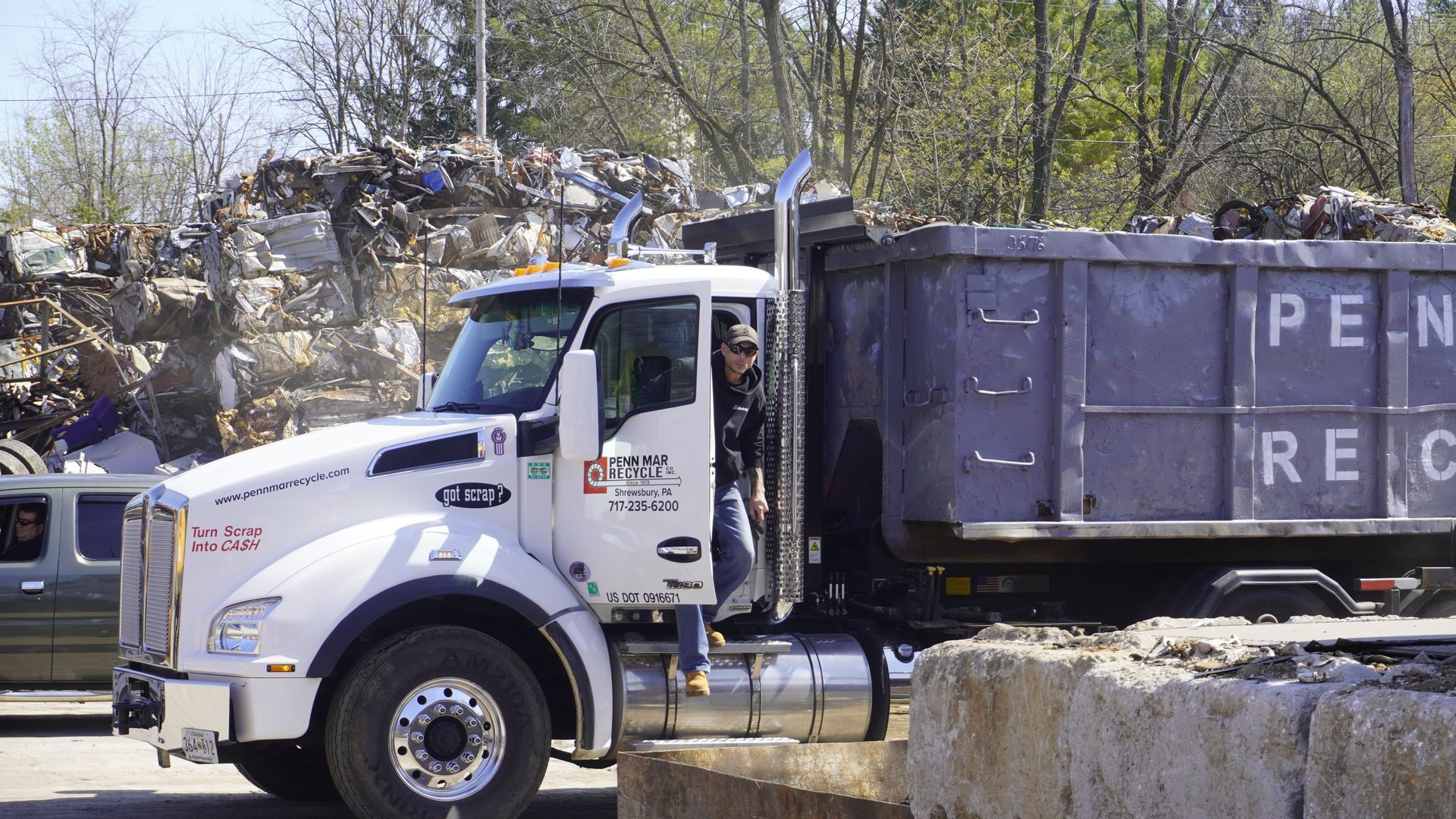 White dump truck with a black container on a road near trees and stone blocks outdoors.