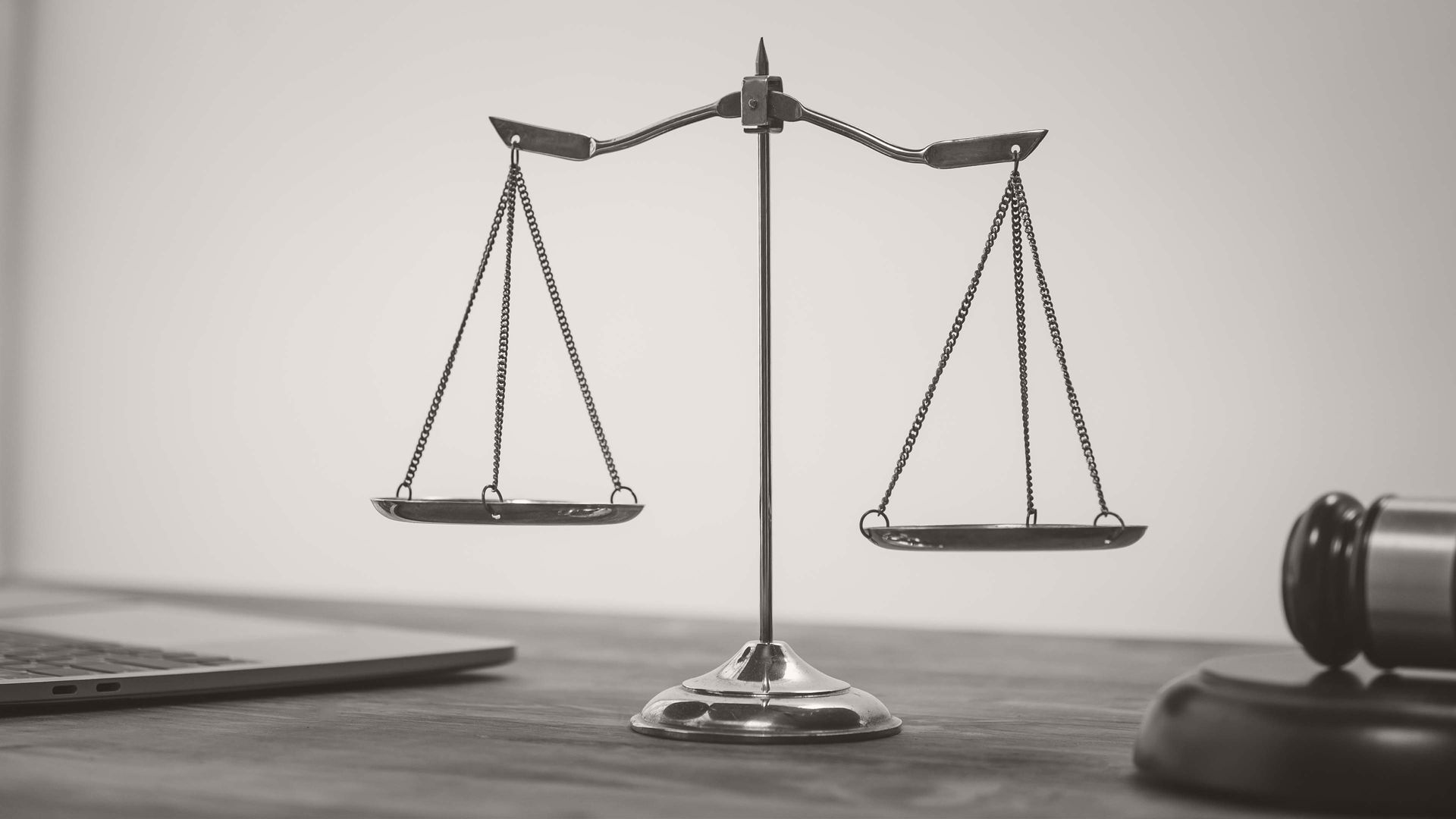 Black-and-white image of a balance scale on a desk, with a laptop and gavel nearby.