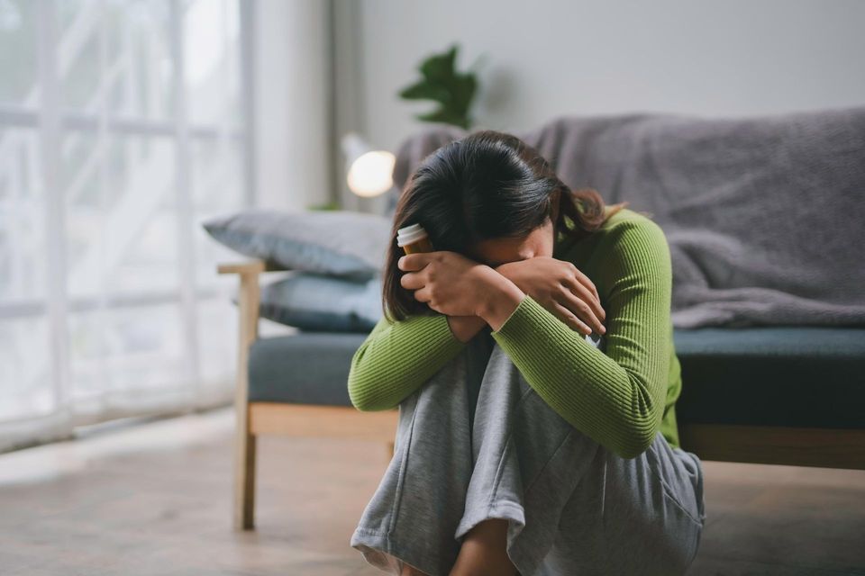 Person sitting on the floor with head in hands beside a couch, appearing upset in a living room.