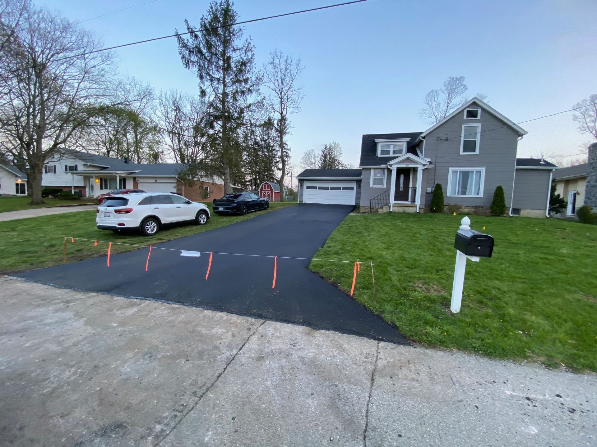 Residential driveway with parked cars, house, and orange stakes marking a newly paved blacktop area.