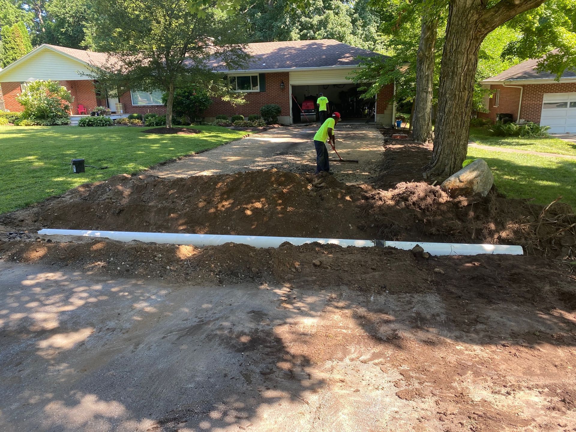 Workers leveling a dirt driveway beside a house, with a white pipe barrier in the foreground.