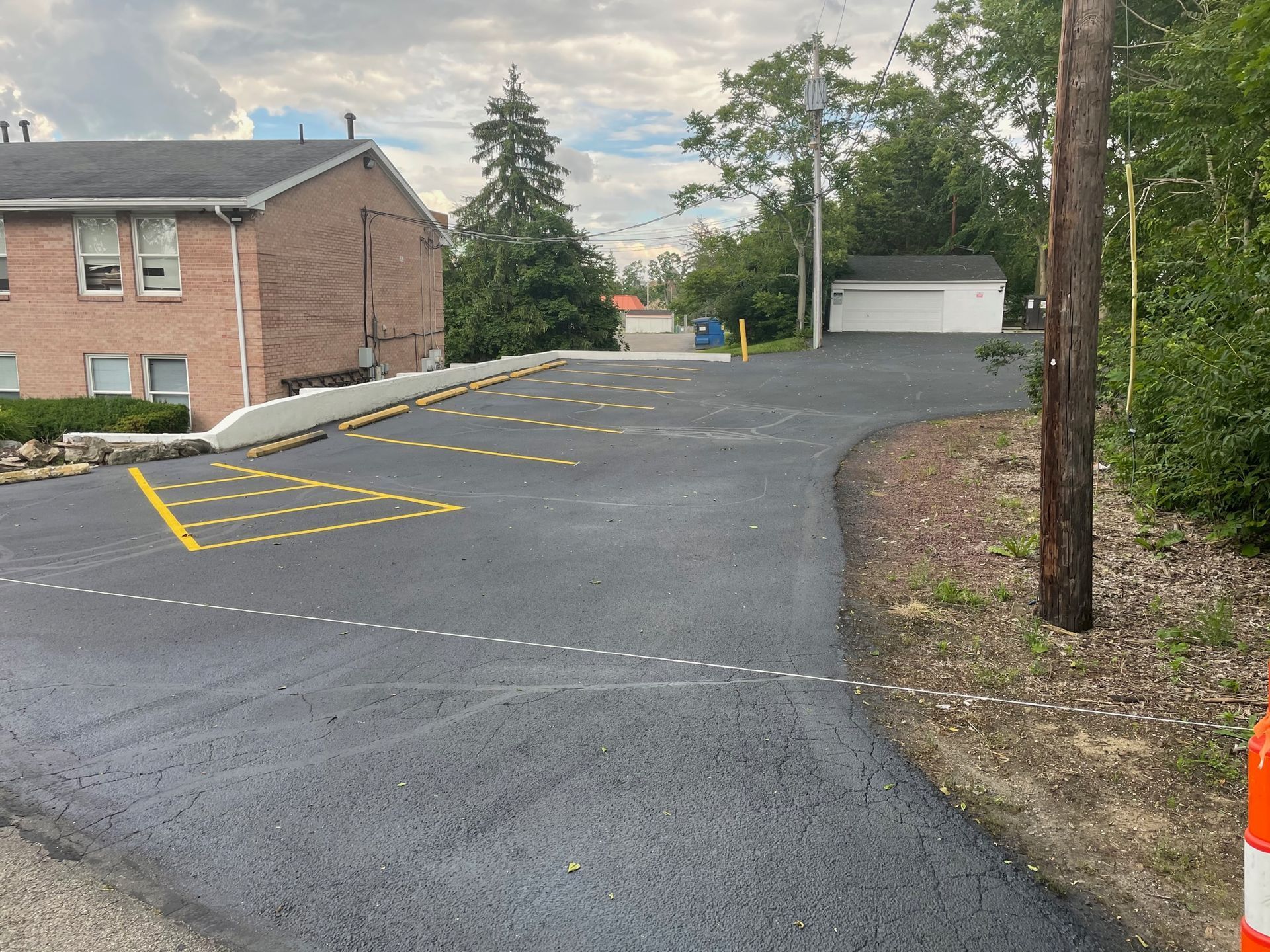 Empty paved parking lot beside a brick building and garage, with yellow marked spaces and trees along the edge.
