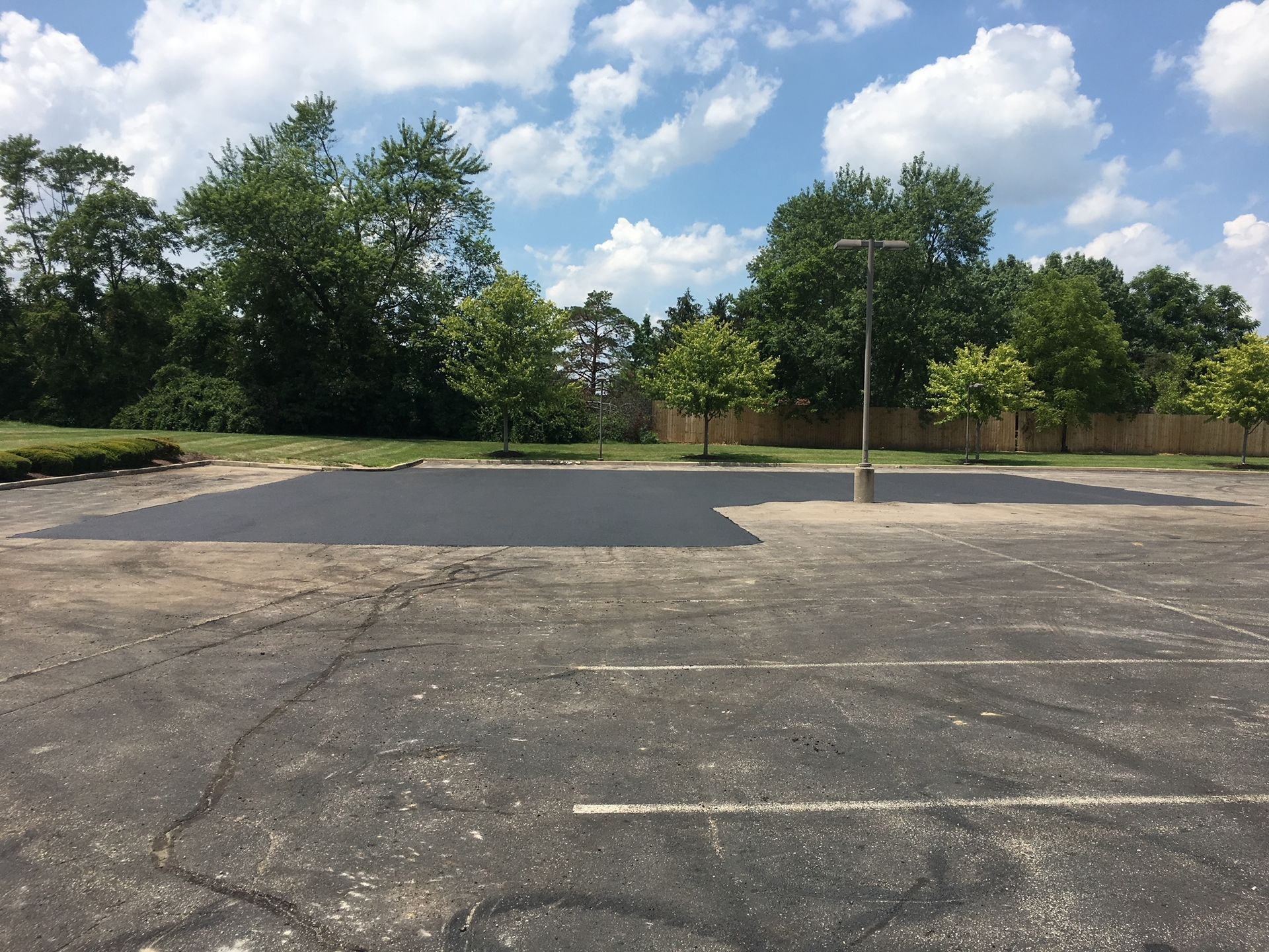 Empty parking lot with cracked asphalt, a tree-lined edge, and blue sky with clouds.
