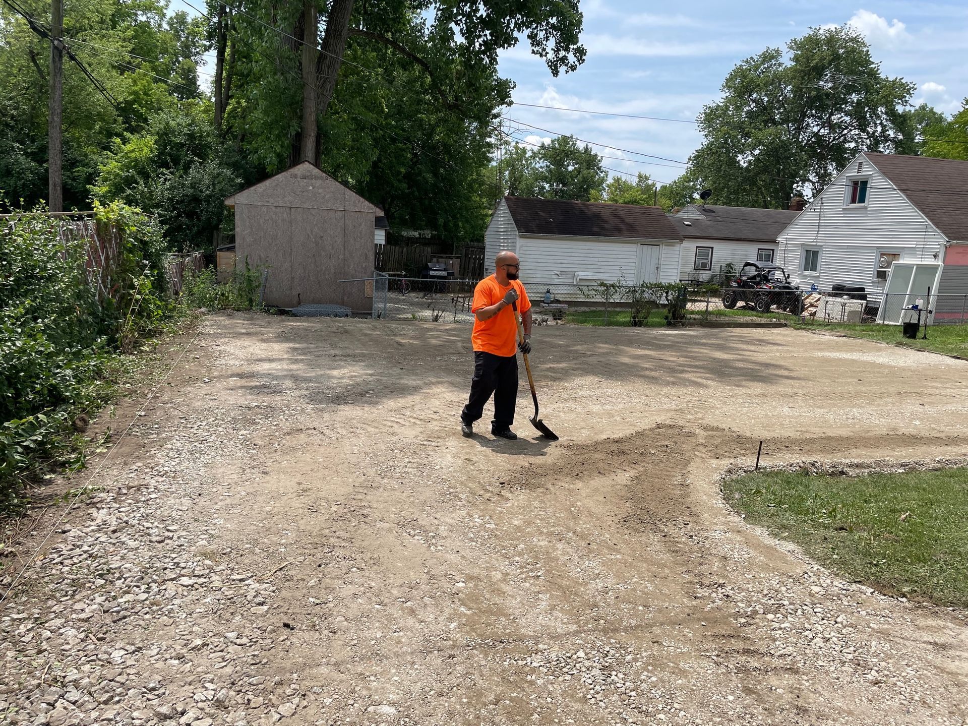 Person in orange shirt raking gravel in a driveway beside houses and trees