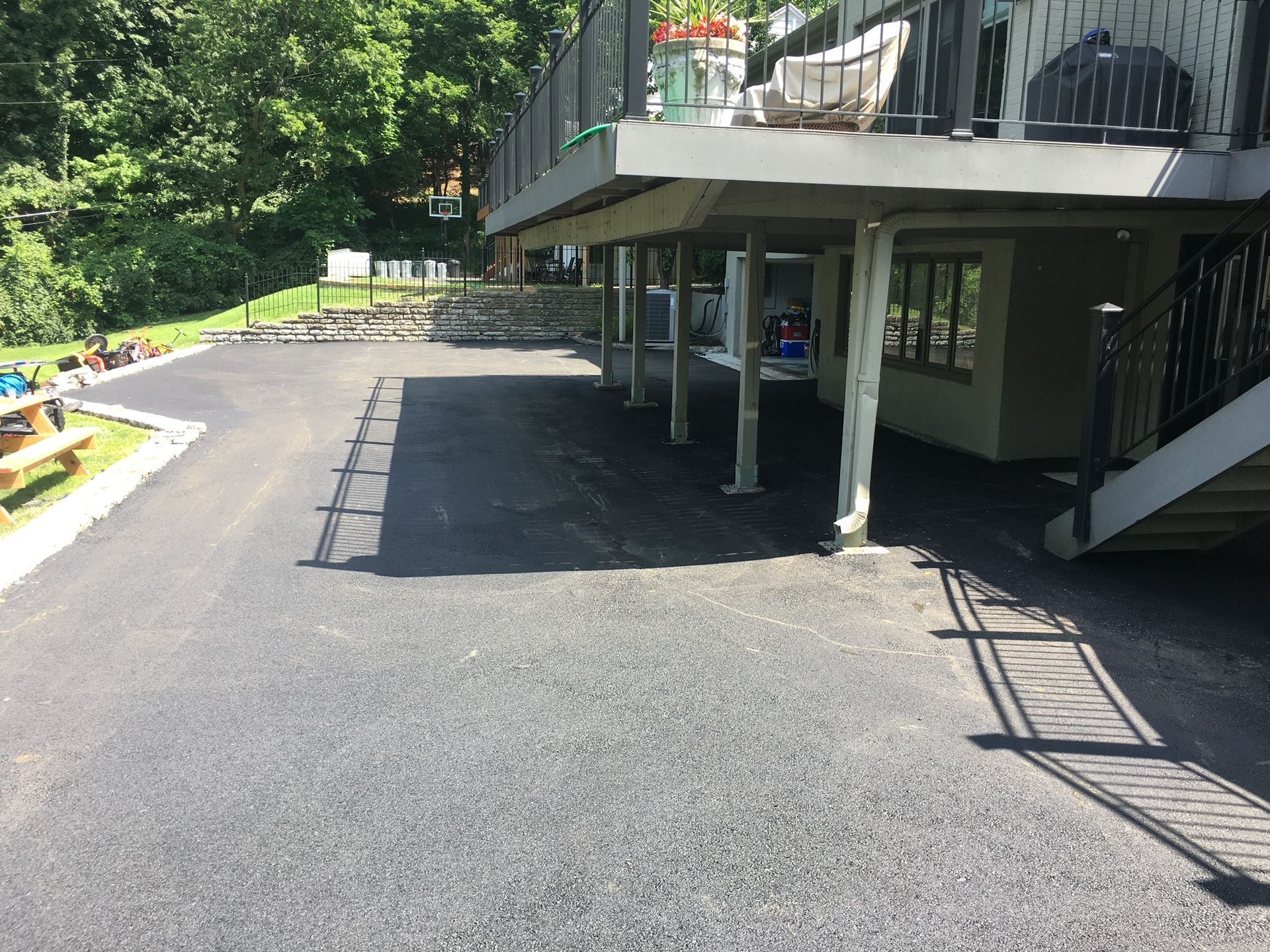 Driveway beside a building with stairs, railings, and a covered patio, bordered by trees and greenery
