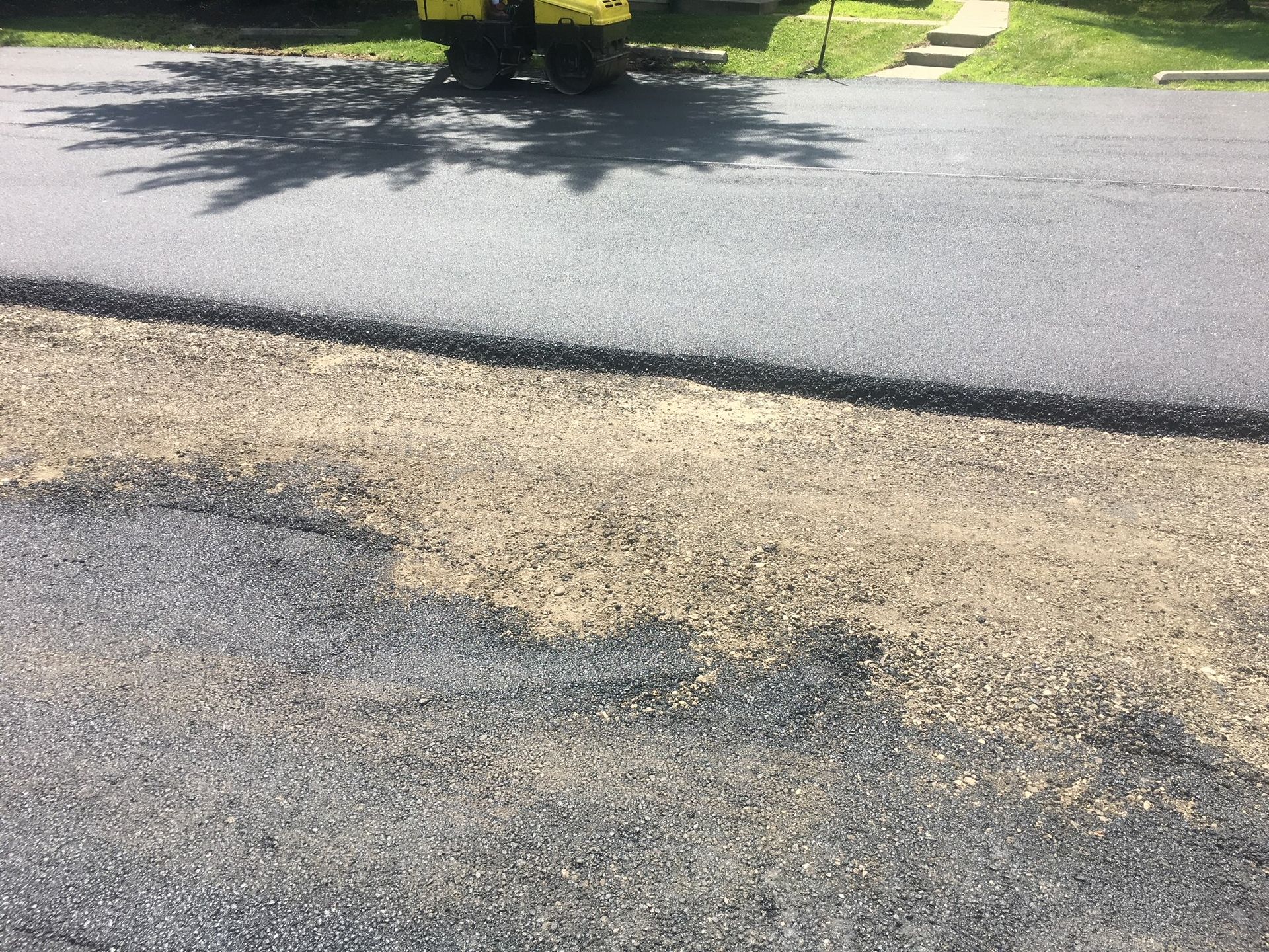 Cracked asphalt road beside a grassy curb in bright sunlight, with tree shadows across the pavement
