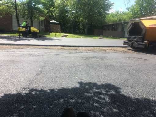 Workers repairing a sunlit residential street beside parked trucks and a roadside building