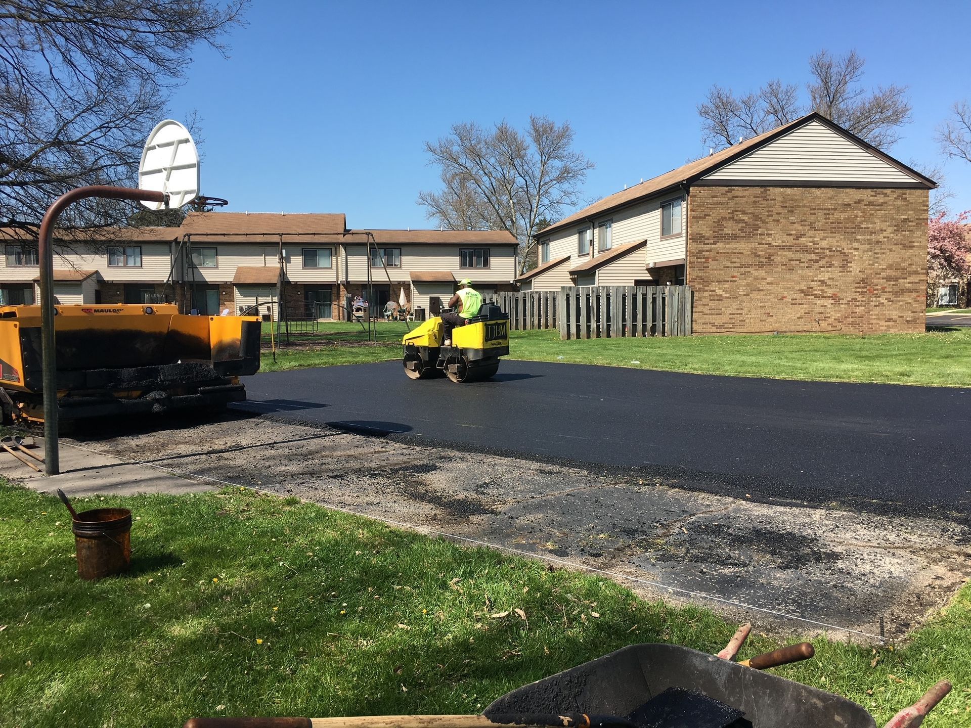 Workers paving a driveway outside brick apartments, with a yellow machine on a sunny day
