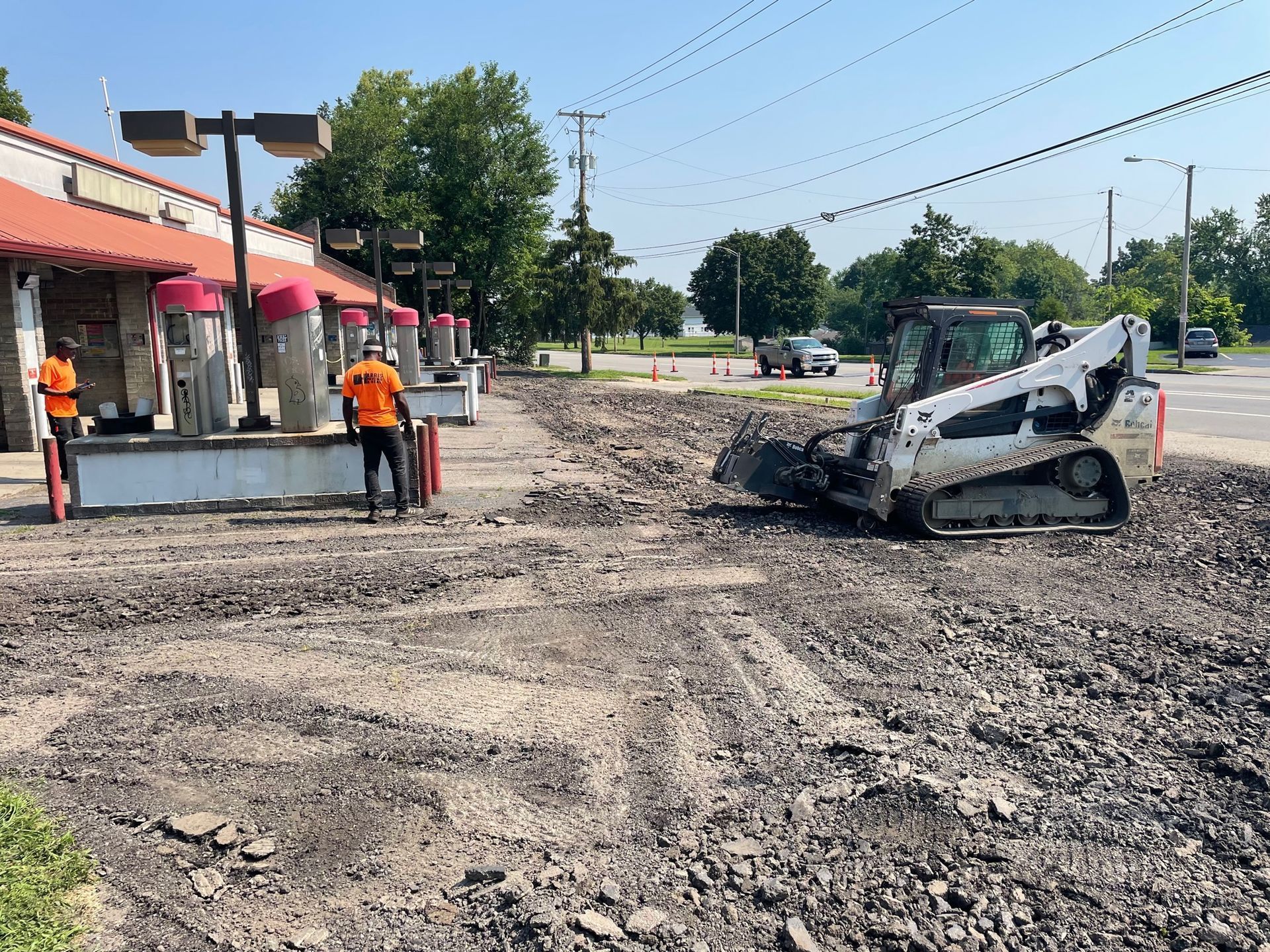 Roadwork scene with a damaged skid steer on a muddy street beside storefronts and workers in orange vests