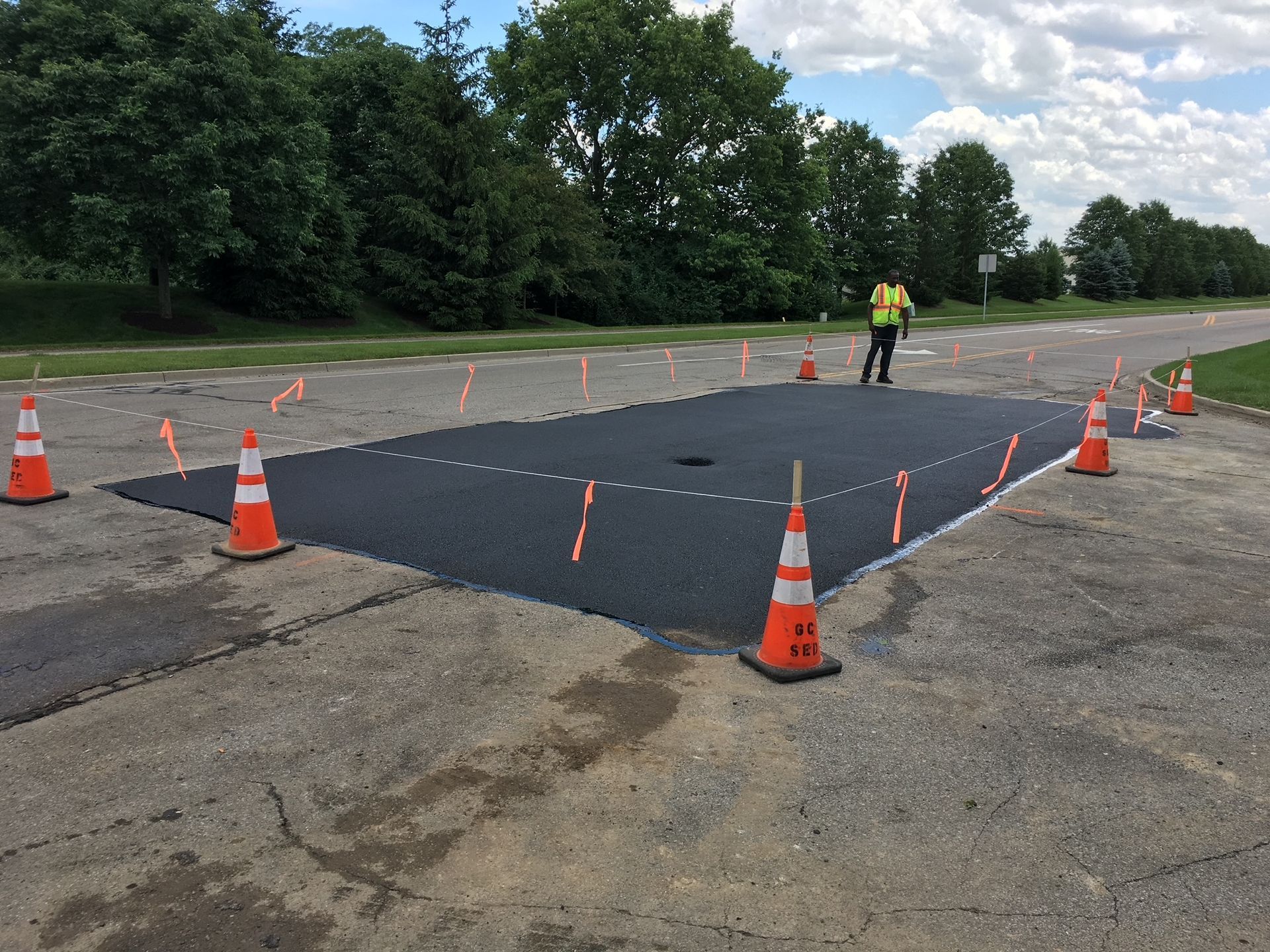 Orange cones and caution tape around a patched asphalt section in a parking lot, with a worker nearby.