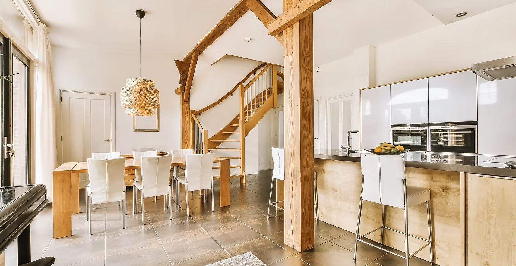 Spacieux séjour ouvert avec table à manger, îlot de cuisine, poutres apparentes et escalier. Tons blancs et bois naturels.