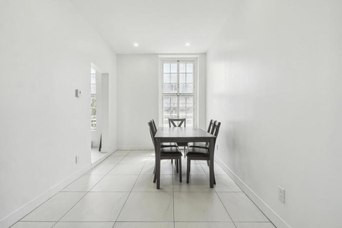 Salle à manger avec table, chaises et fenêtre grises. Murs blancs et carrelage au sol.