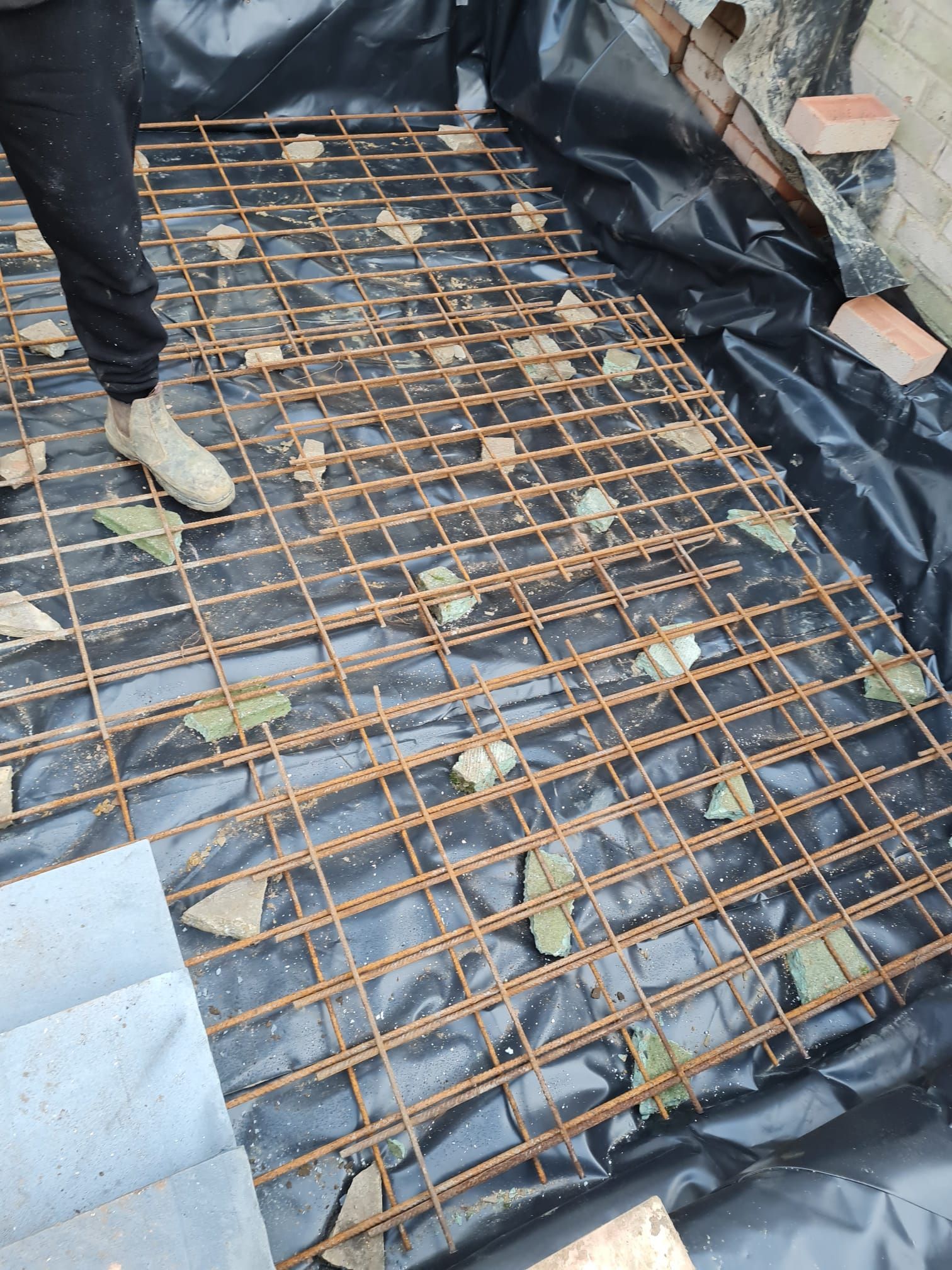Steel rebar grid laid over black plastic, partially covering white fabric, with leaves scattered. Person's legs visible.