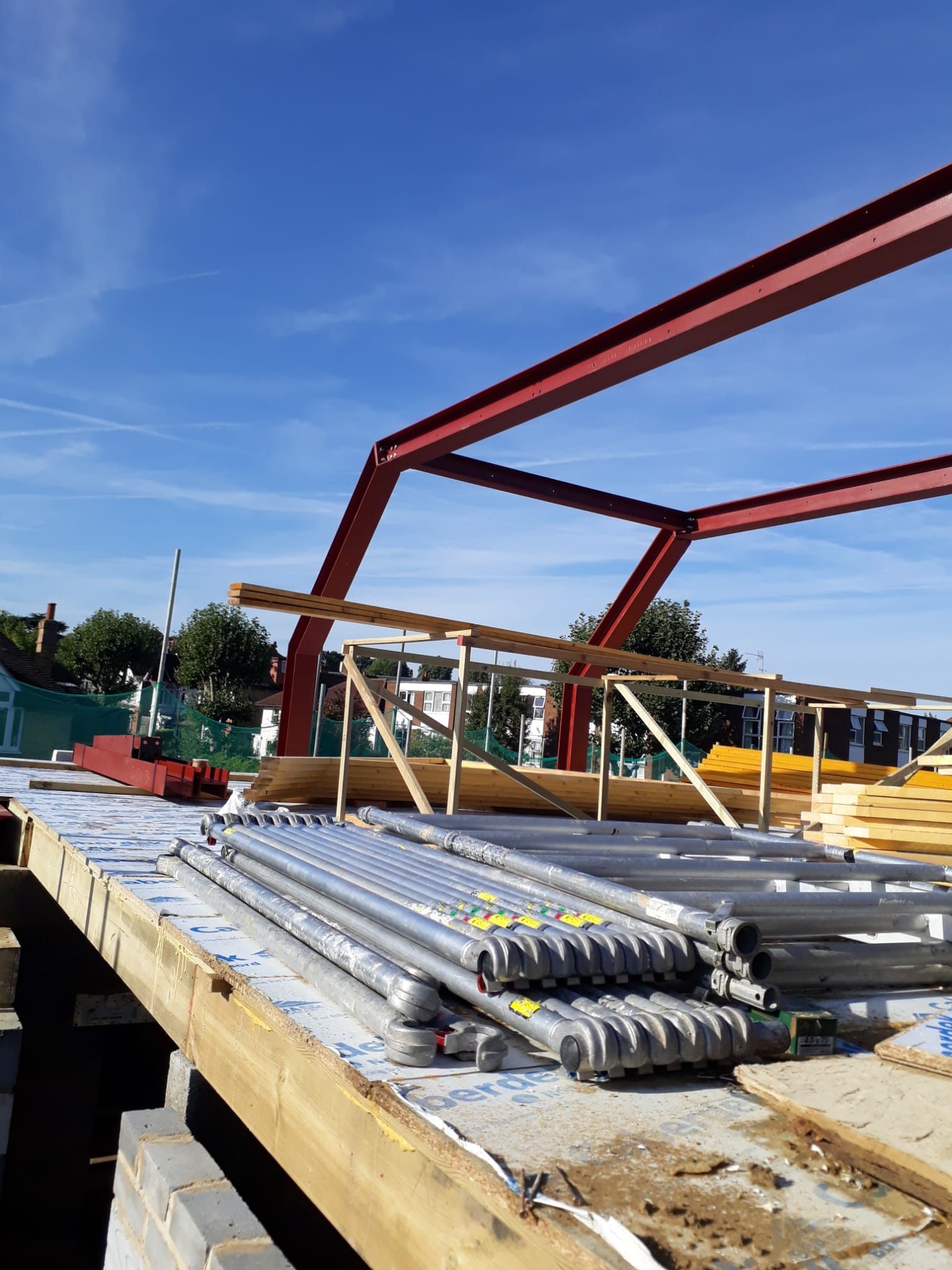 Construction site with red steel frame, scaffolding, and lumber under a blue sky.