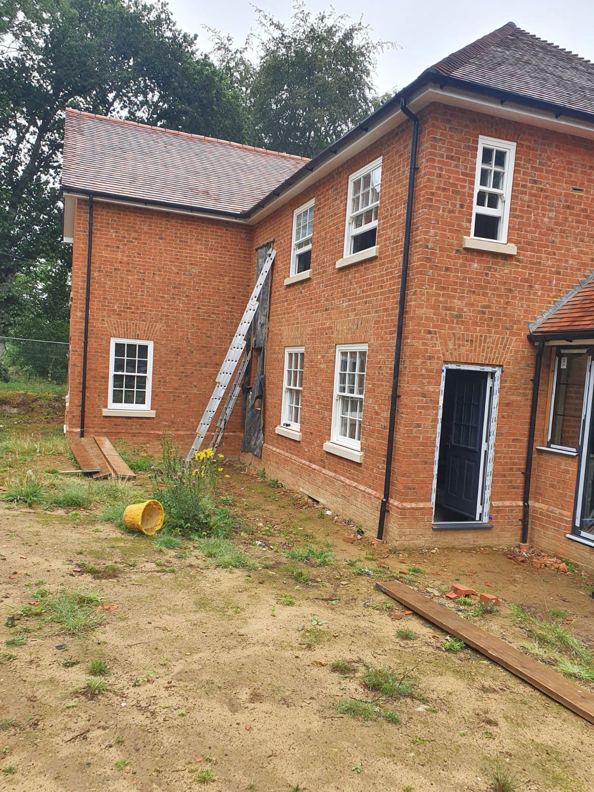 Red brick house under construction with a ladder leaning against it. Cloudy day, dirt yard.