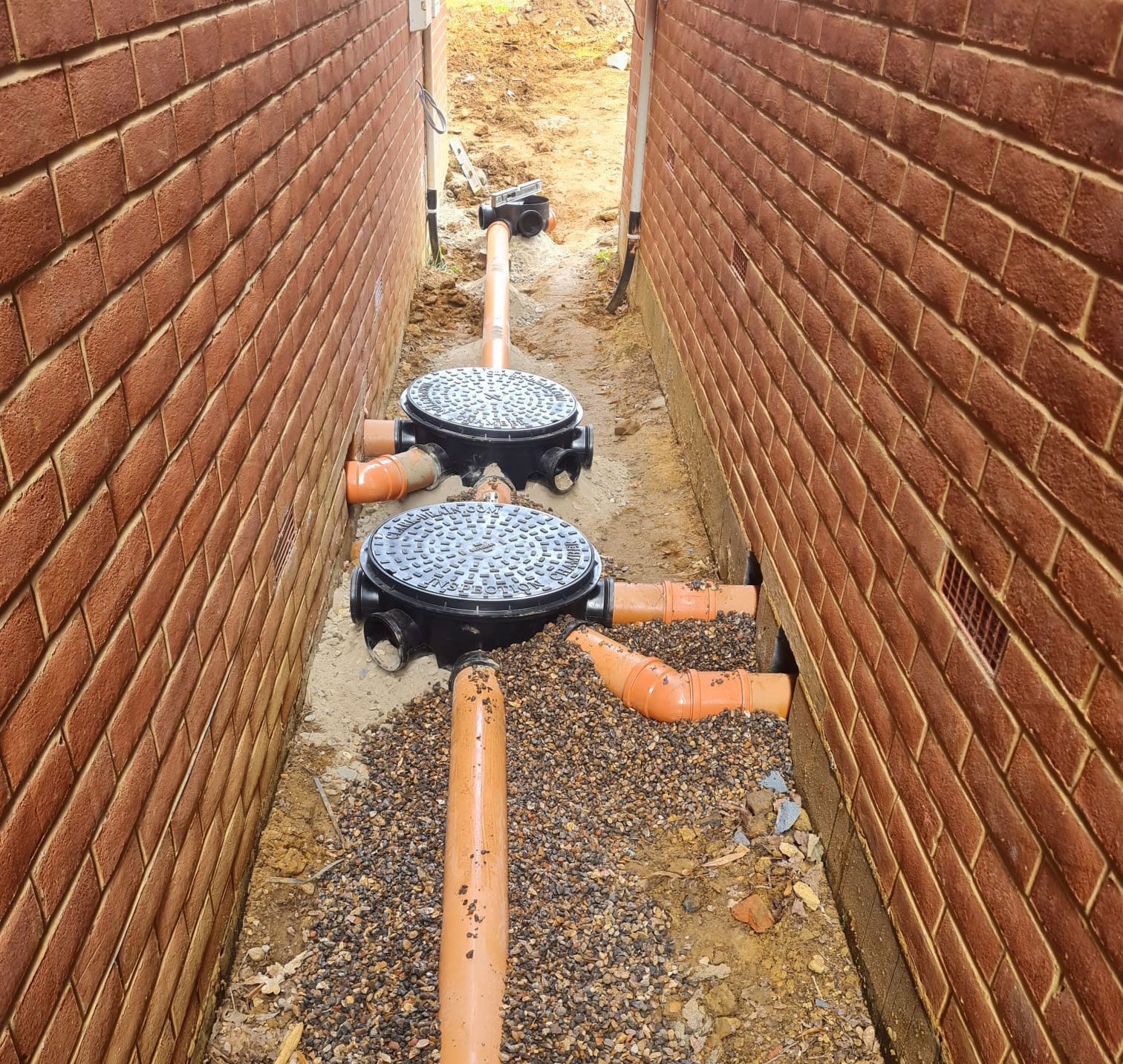 Narrow trench between brick walls with drainage pipes and two black basins.