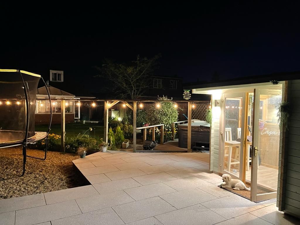 Backyard at night, lit by string lights. A dog rests by a brightly lit shed, with a trampoline to the left.