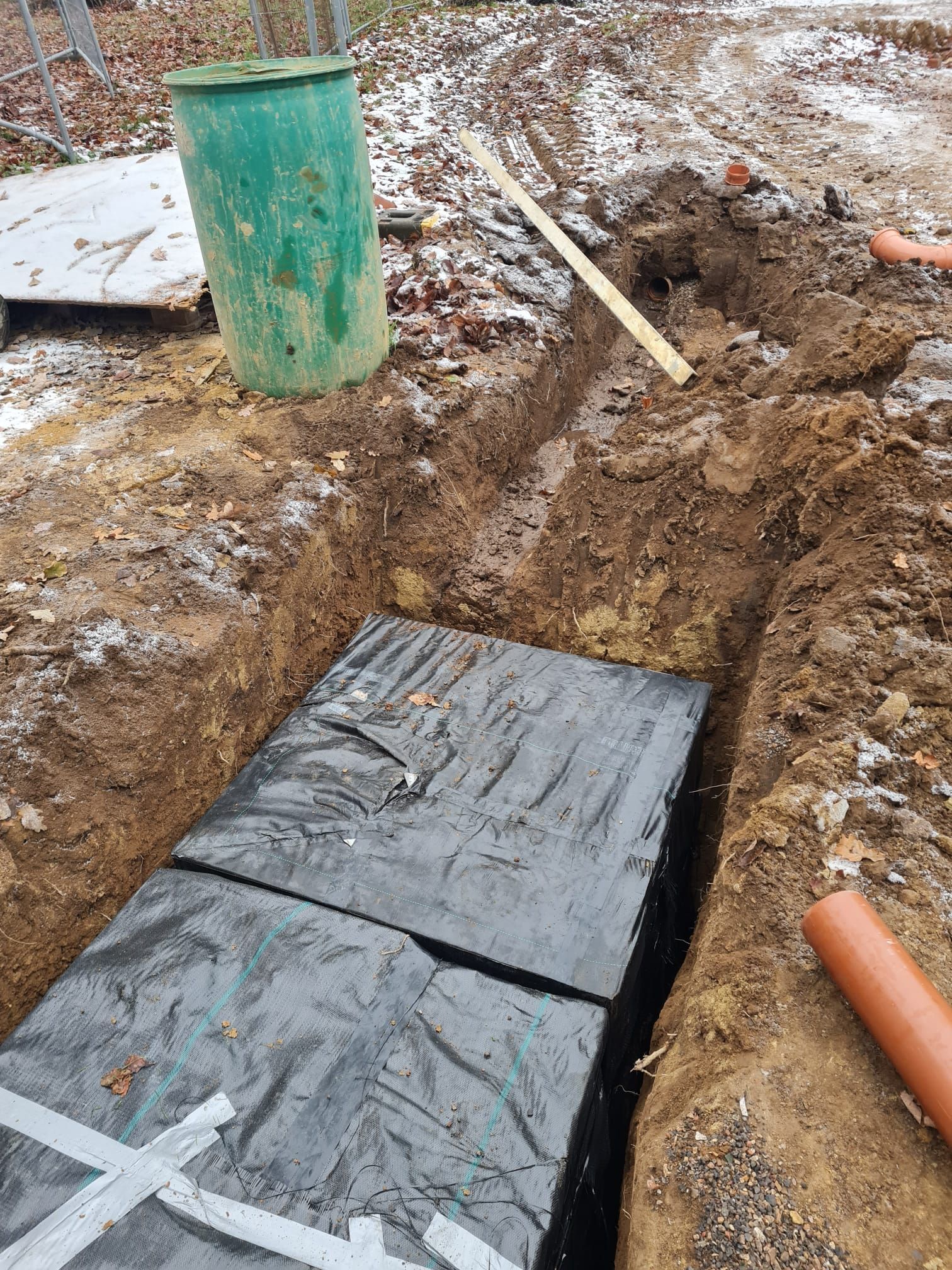A rectangular trench dug in the ground, covered with black material. A green barrel and brown pipe nearby.