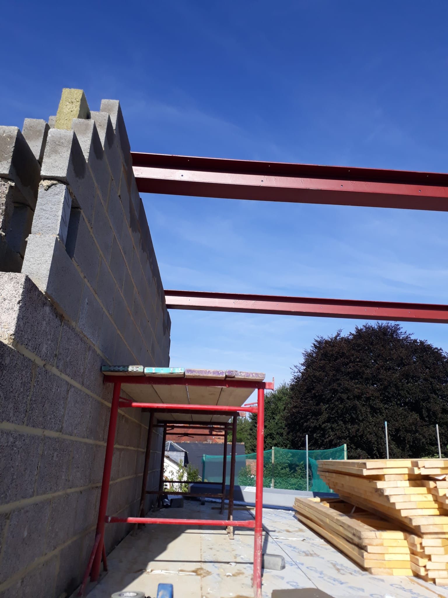 Construction site: Block wall with red steel beams, scaffolding, and stacked lumber under a blue sky.