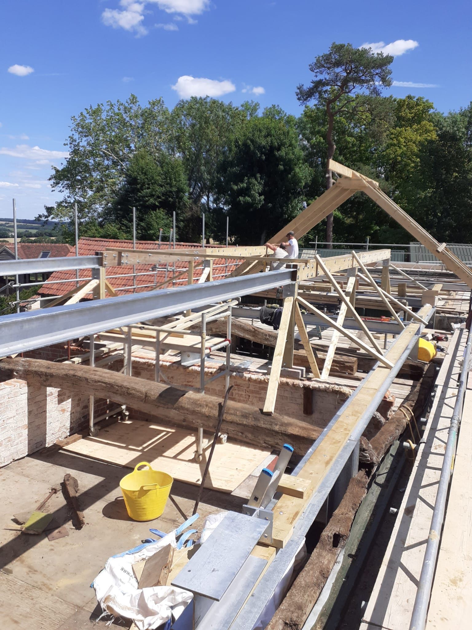 Construction site: partially built roof frame with steel beams, wooden supports, and brick walls under a blue sky.