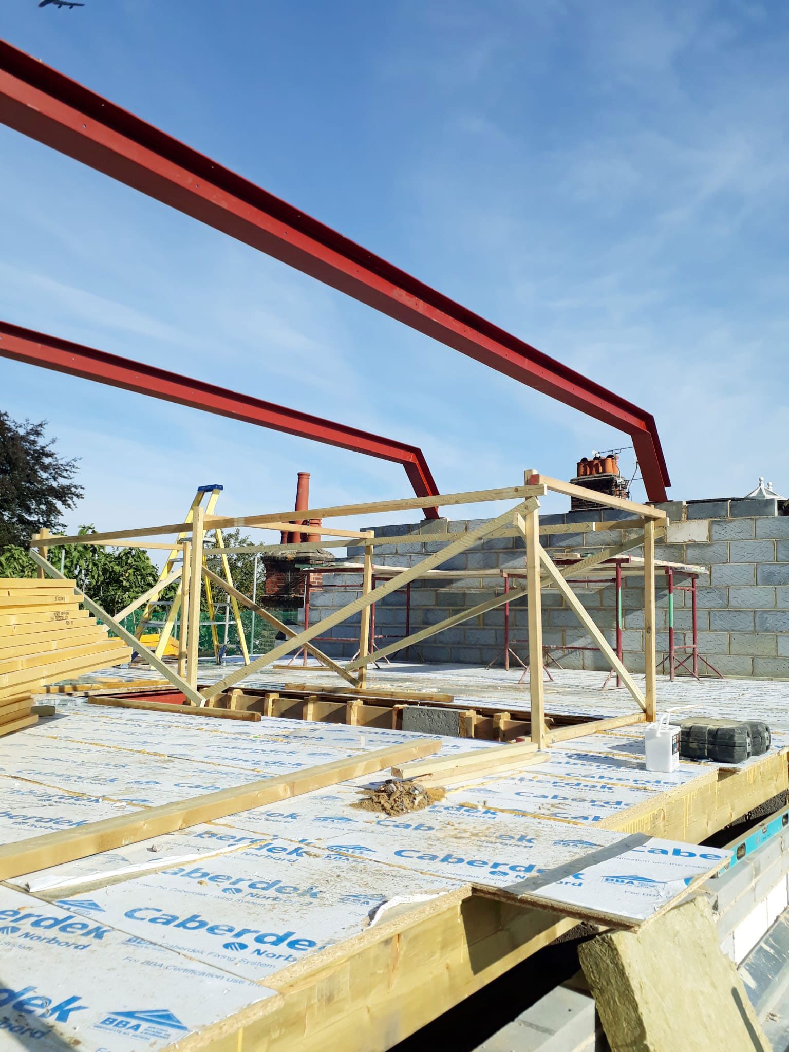 Construction site with exposed wooden framing and red steel beams against a blue sky.