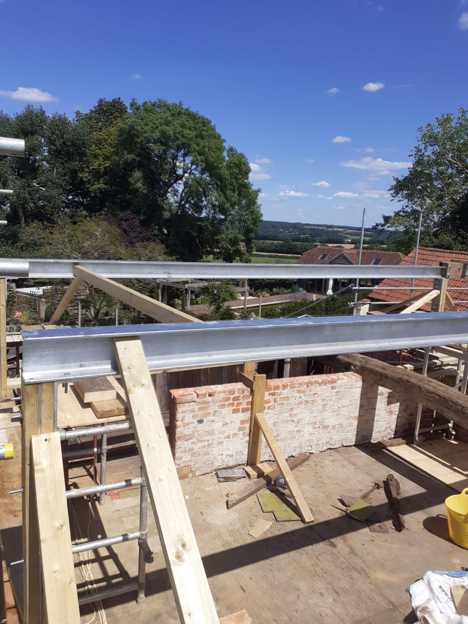 Construction site: steel beams, brick walls, wood supports, and scaffolding, under a blue sky.