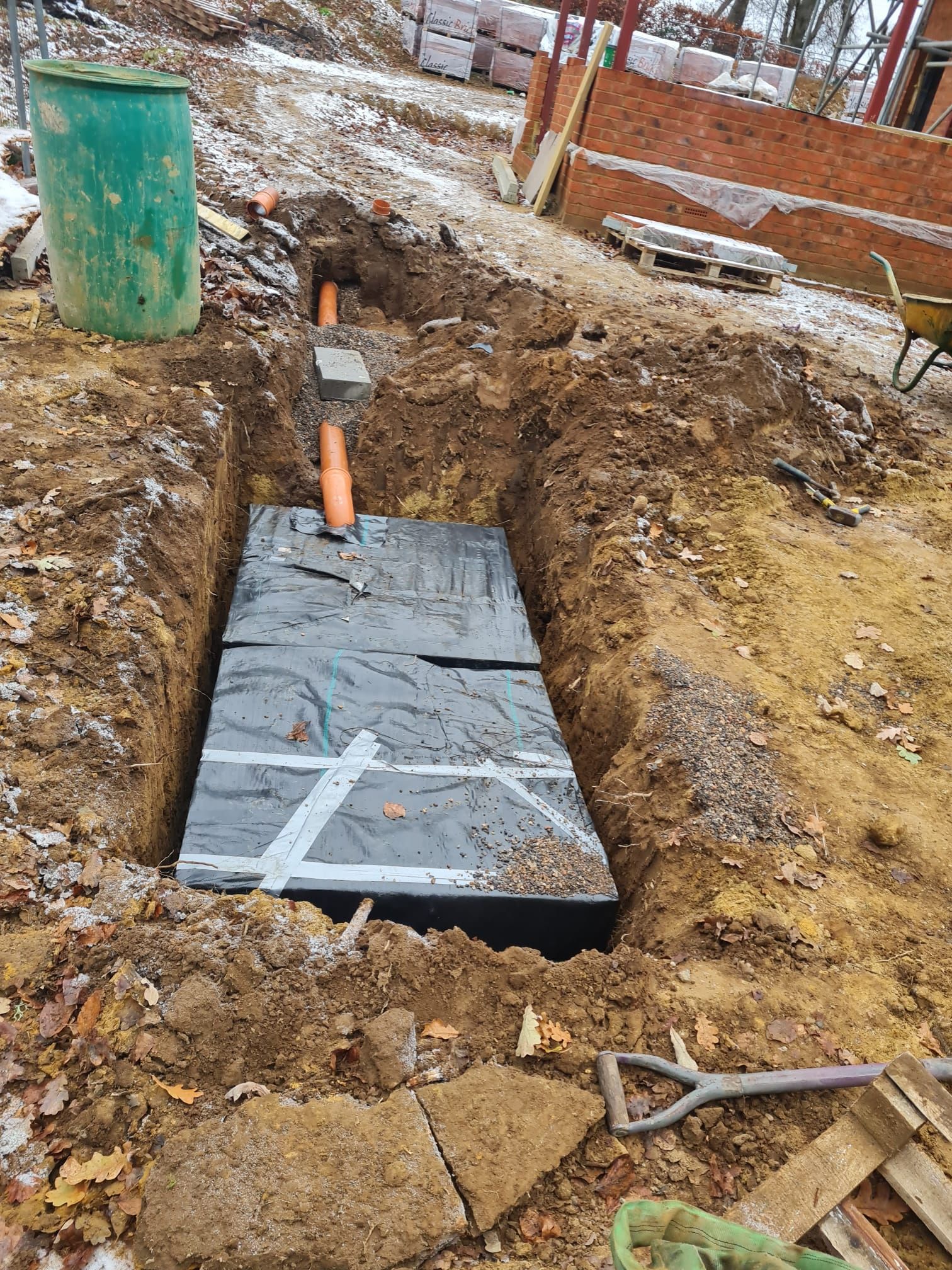 Open excavation with a buried, covered rectangular structure, orange pipe, and green barrel. Construction site.