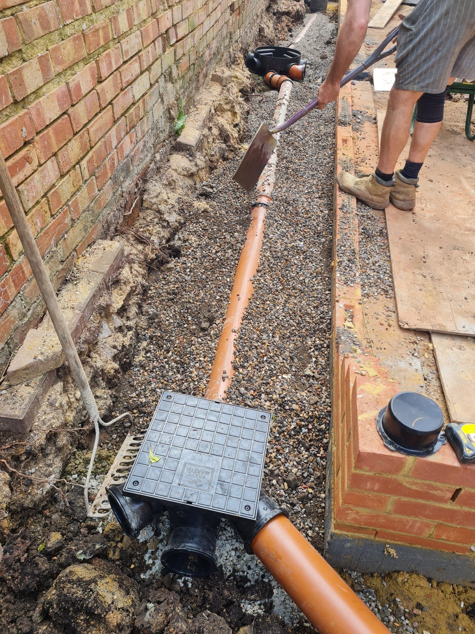 Man shoveling gravel into a trench with drainage pipes next to a brick wall and patio.