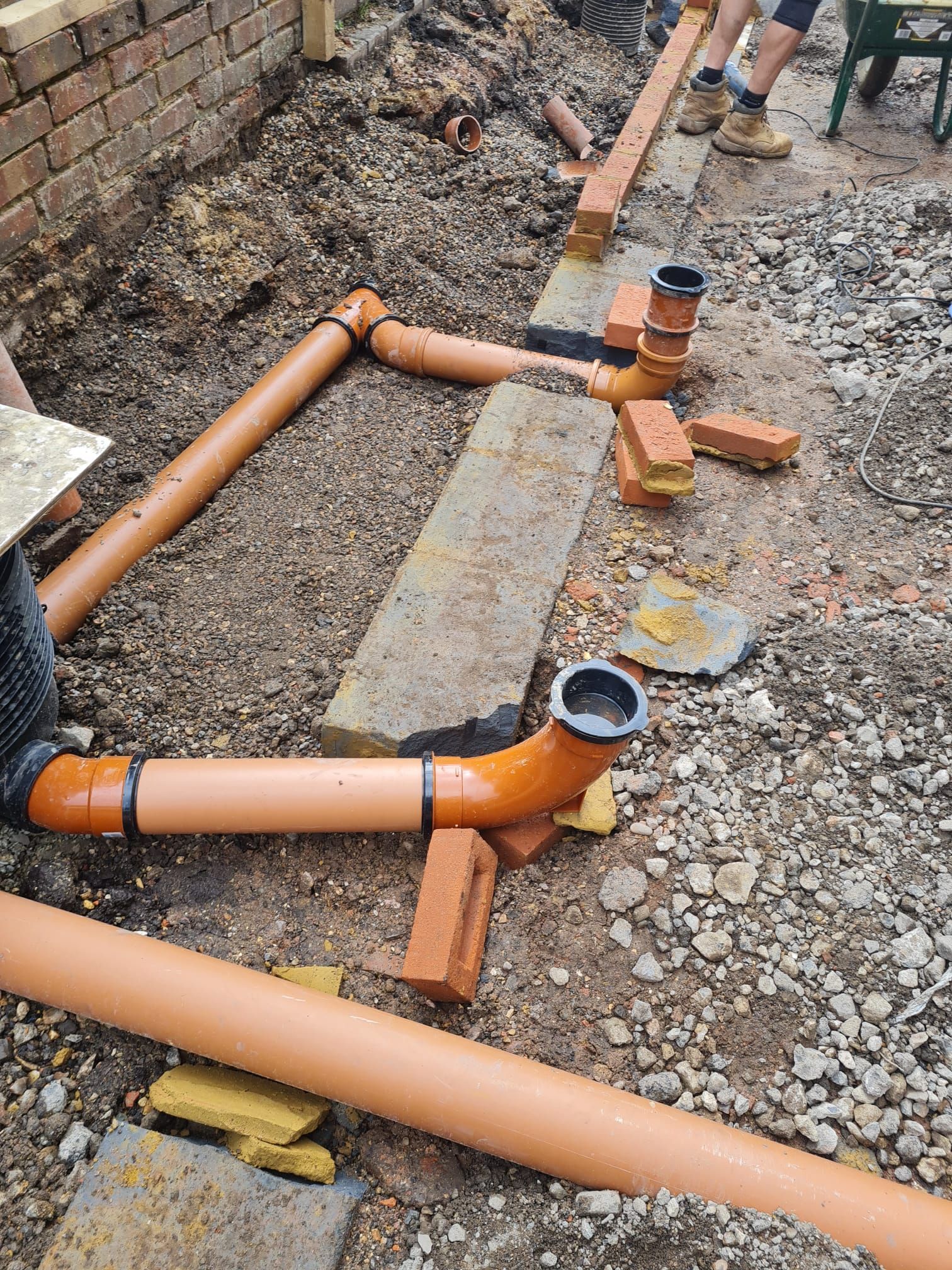 Orange plastic pipes and fittings being installed in a trench next to a brick wall.