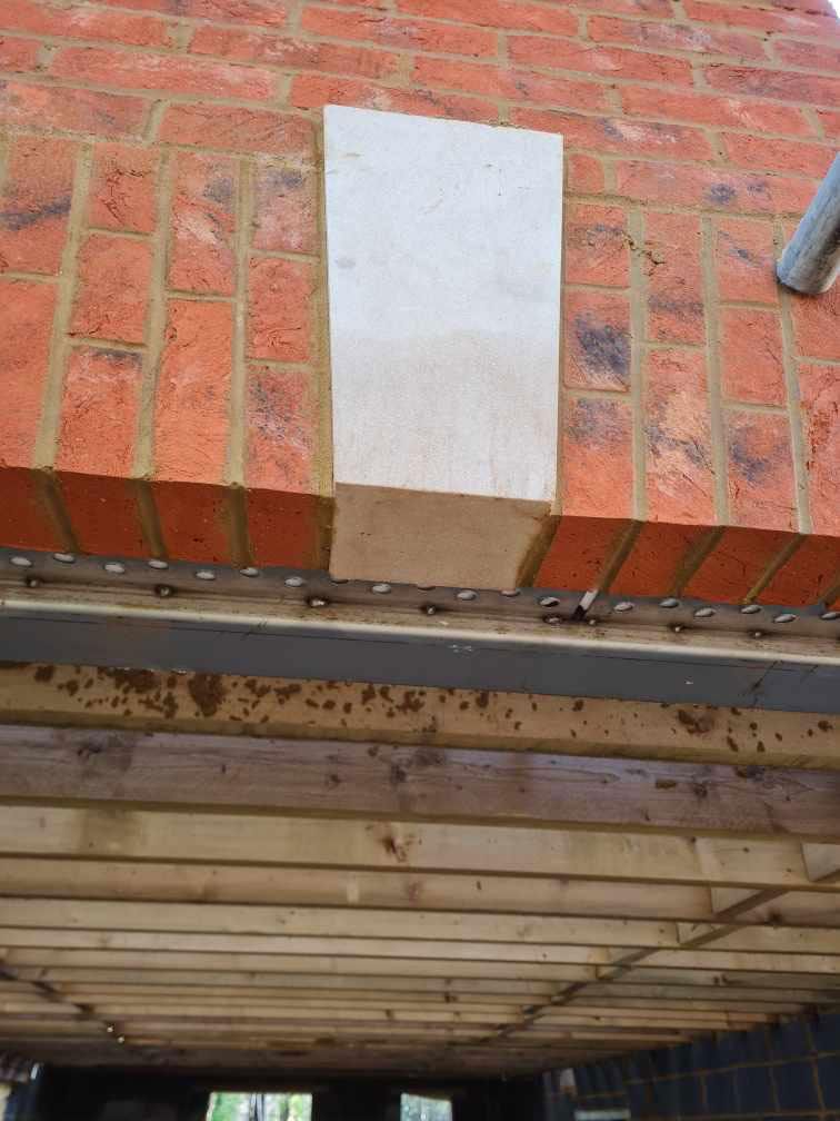 Brick building under construction, with a rectangular stone panel partially installed. Wooden beams visible below.
