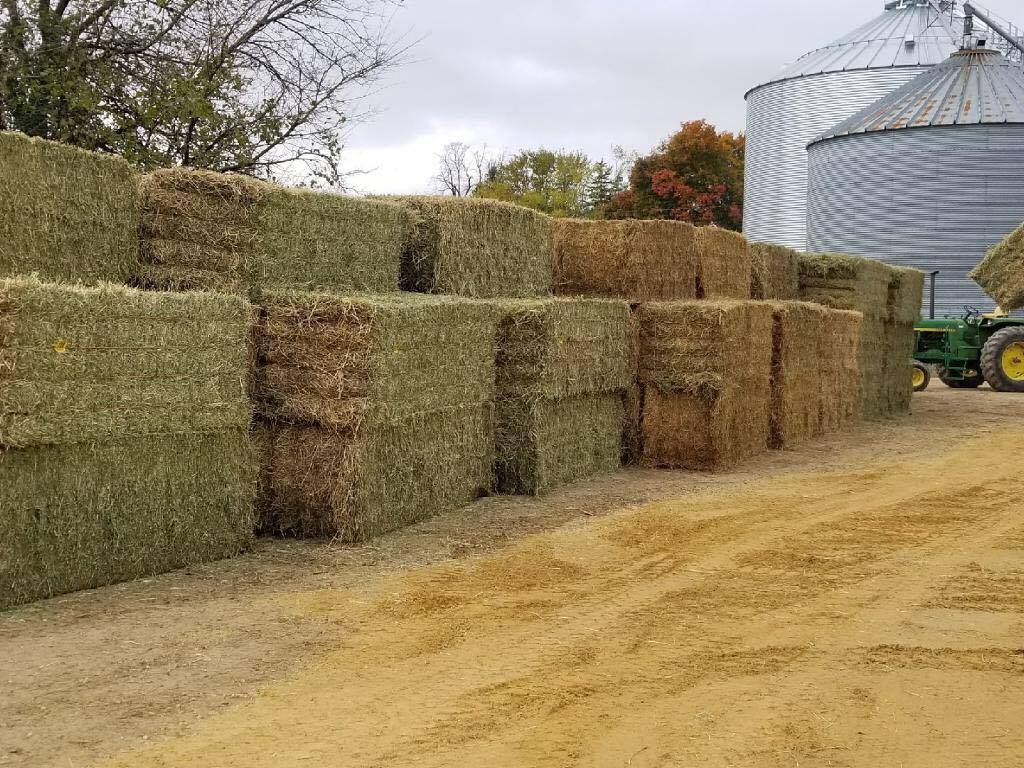 Stacked hay bales beside a dirt road near a grain silo and tractor.