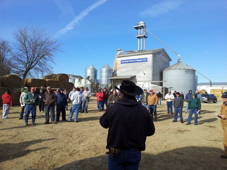 Tour group gathered outside a grain elevator on a sunny farm, facing a guide in a black hat