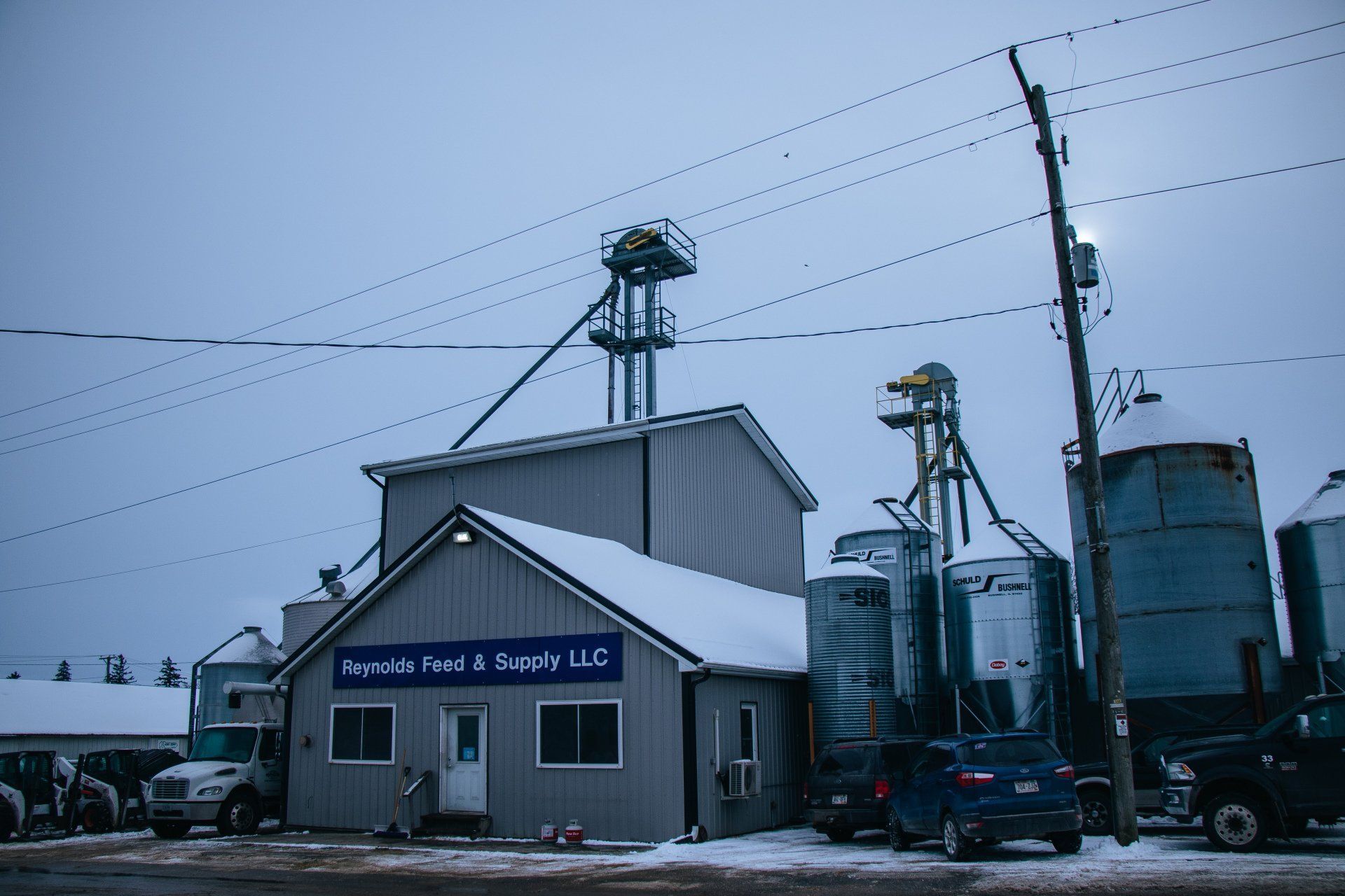 Snowy grain elevator and farm buildings under a gray sky, with power lines overhead and a truck parked nearby
