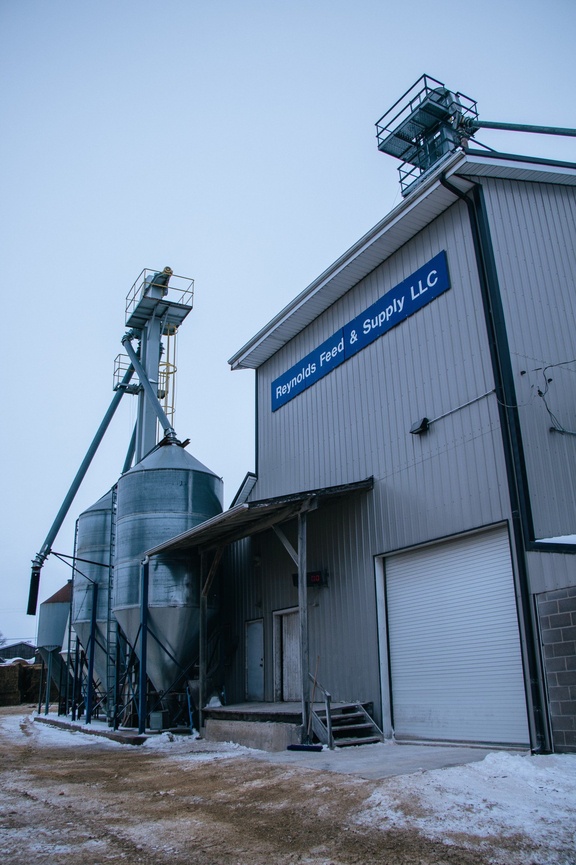 Snowy industrial grain elevator beside a warehouse with metal silos and a blue sign