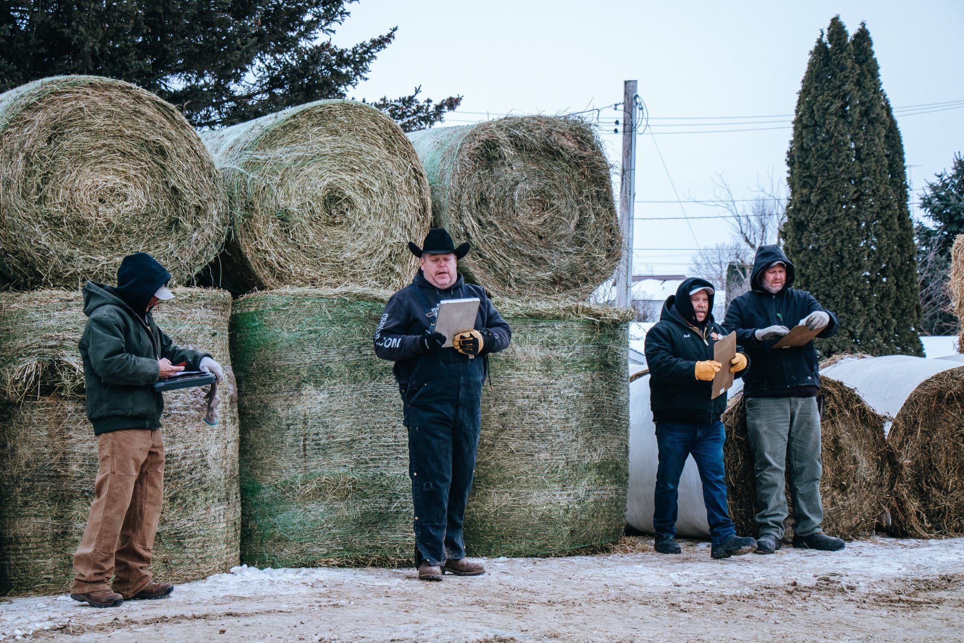 Four people stand beside stacked hay bales in a snowy field, reading papers outdoors.