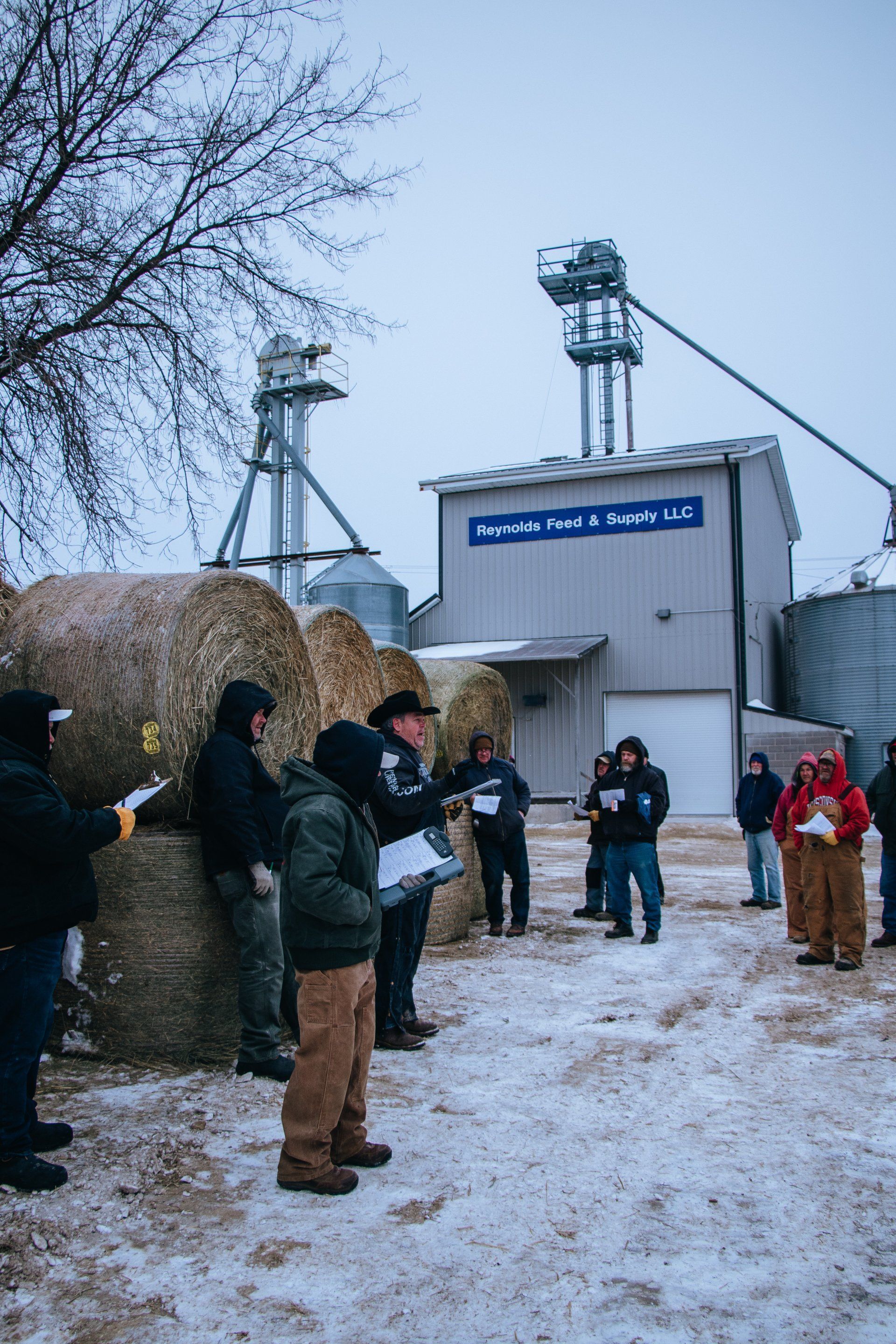 People gathered outside a snowy industrial building, some reading from papers near a hay bale wall.