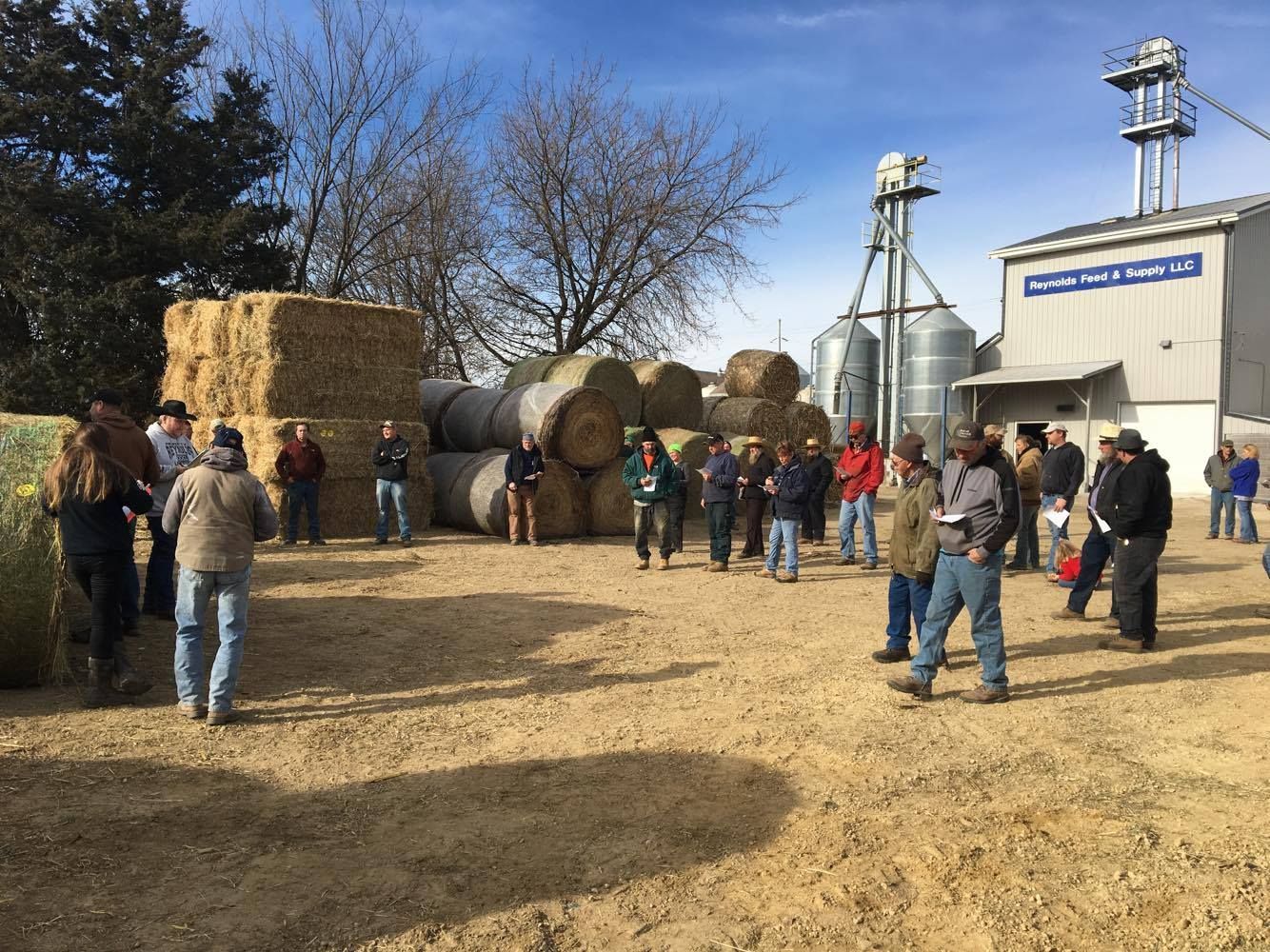 People gathered around hay bales and farm equipment outside a white building on a sunny day