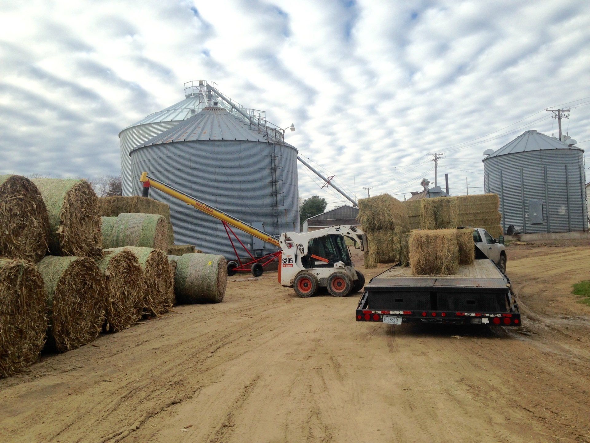 Farmyard with hay bales, a skid steer, grain silos, and a trailer under a cloudy sky.