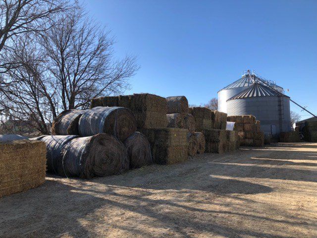 Hay bales and wrapped silage stacked beside a silver grain silo on a farm under a clear blue sky