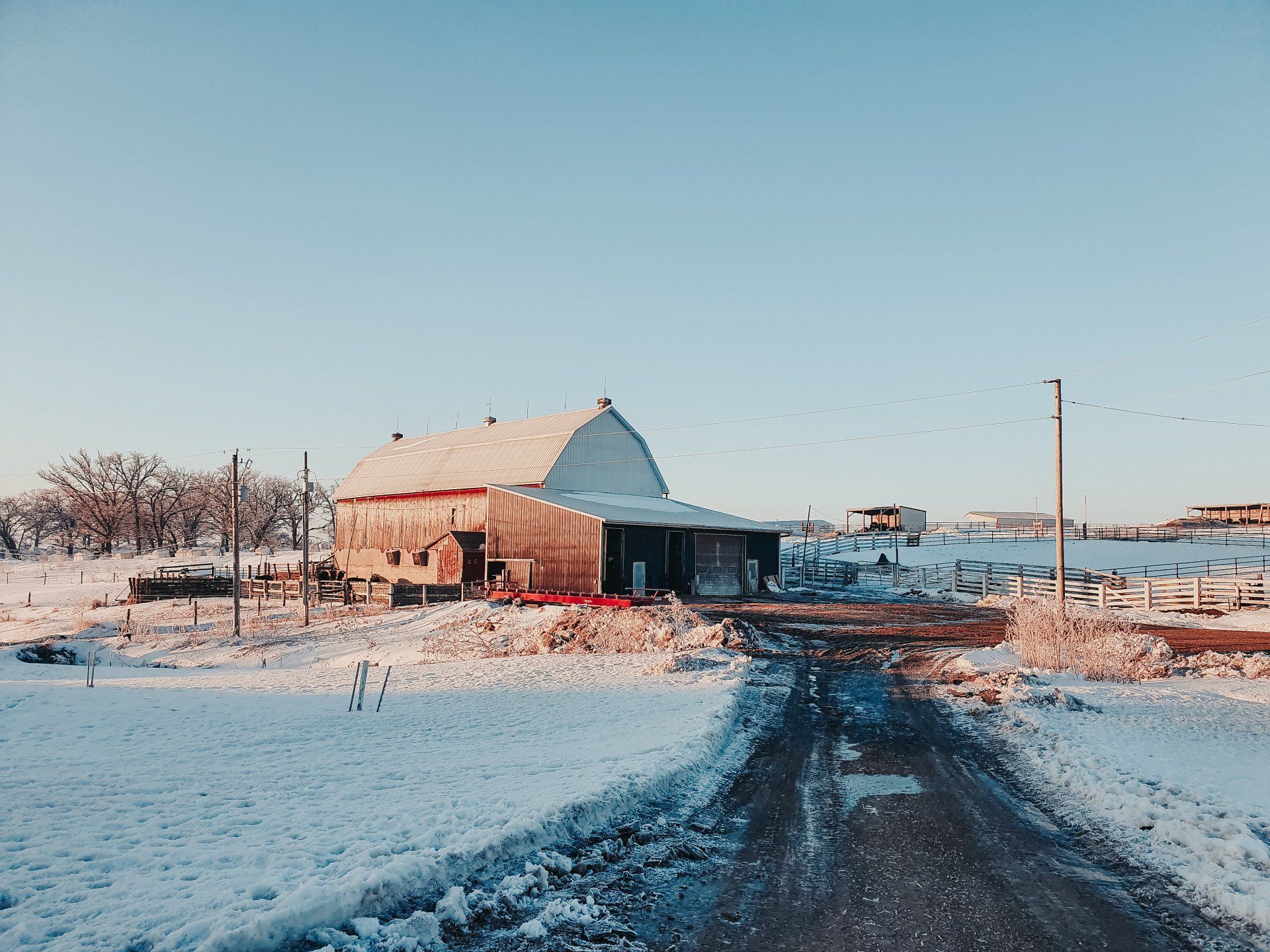 Snowy rural house and driveway under a pale blue sky, with a red barn and fence in the distance