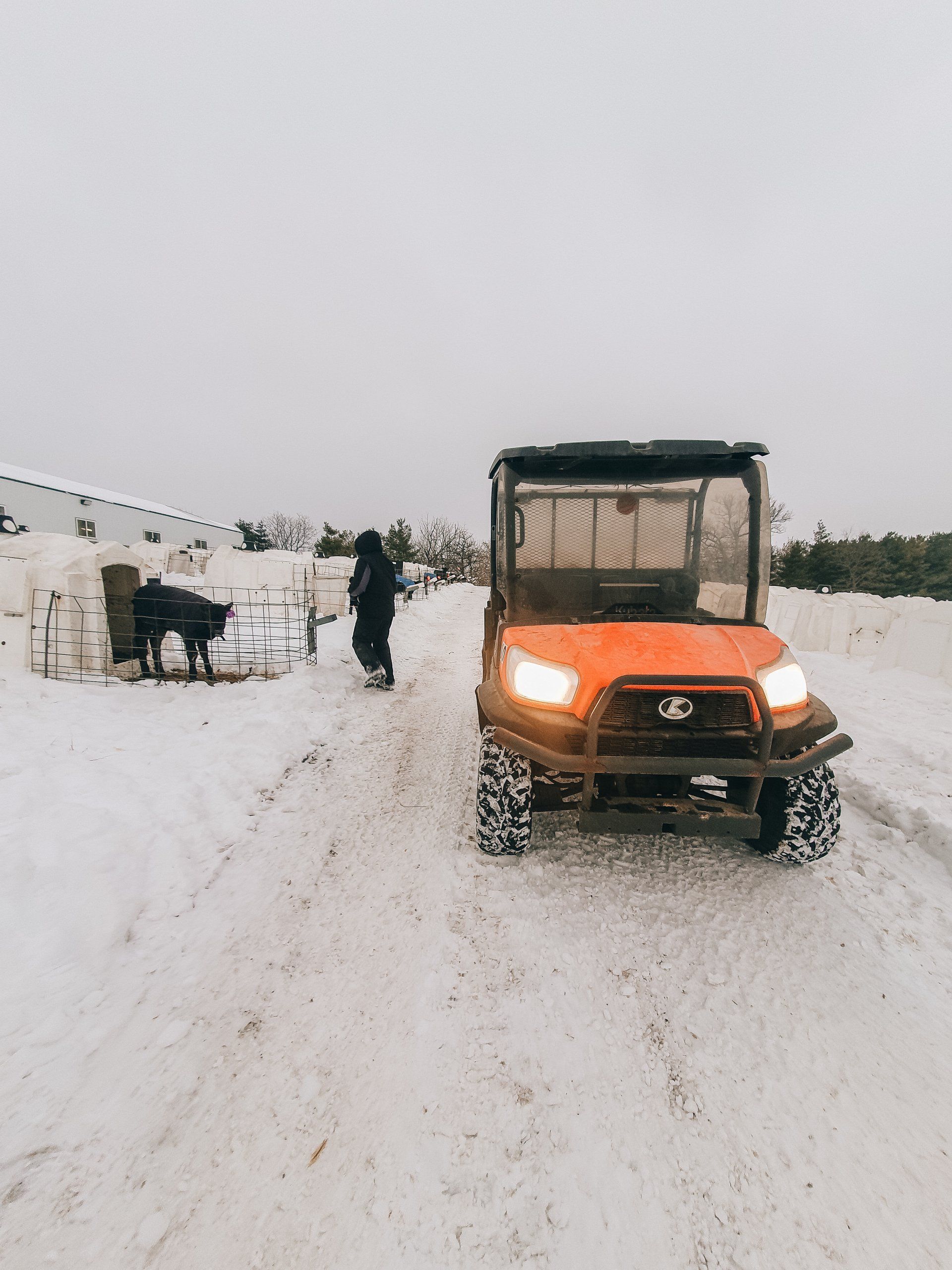 Orange utility vehicle driving on a snowy road beside fences, with a person standing nearby.