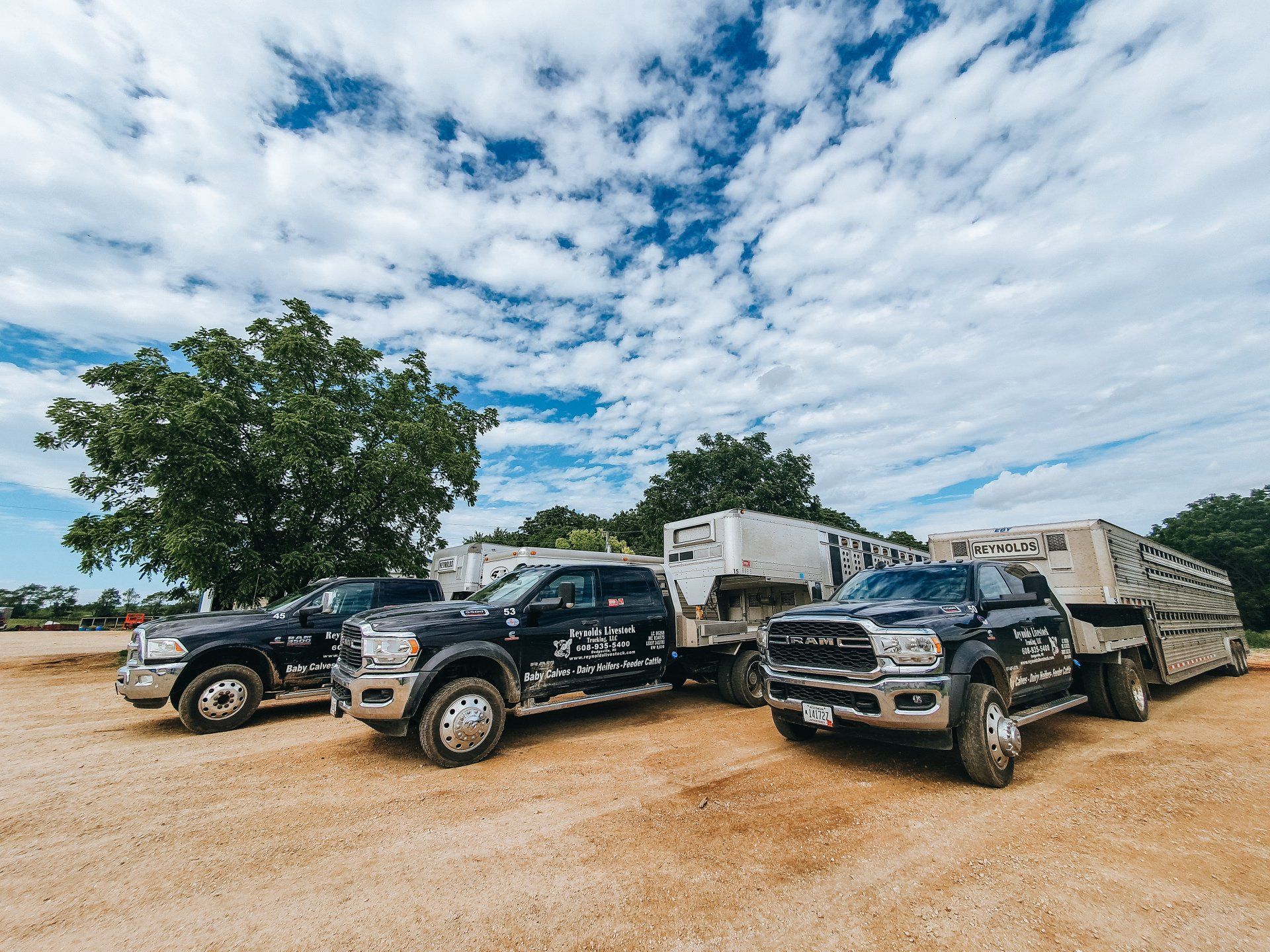 Three black off-road trucks parked on a dirt lot under a cloudy sky, with trailers behind them.