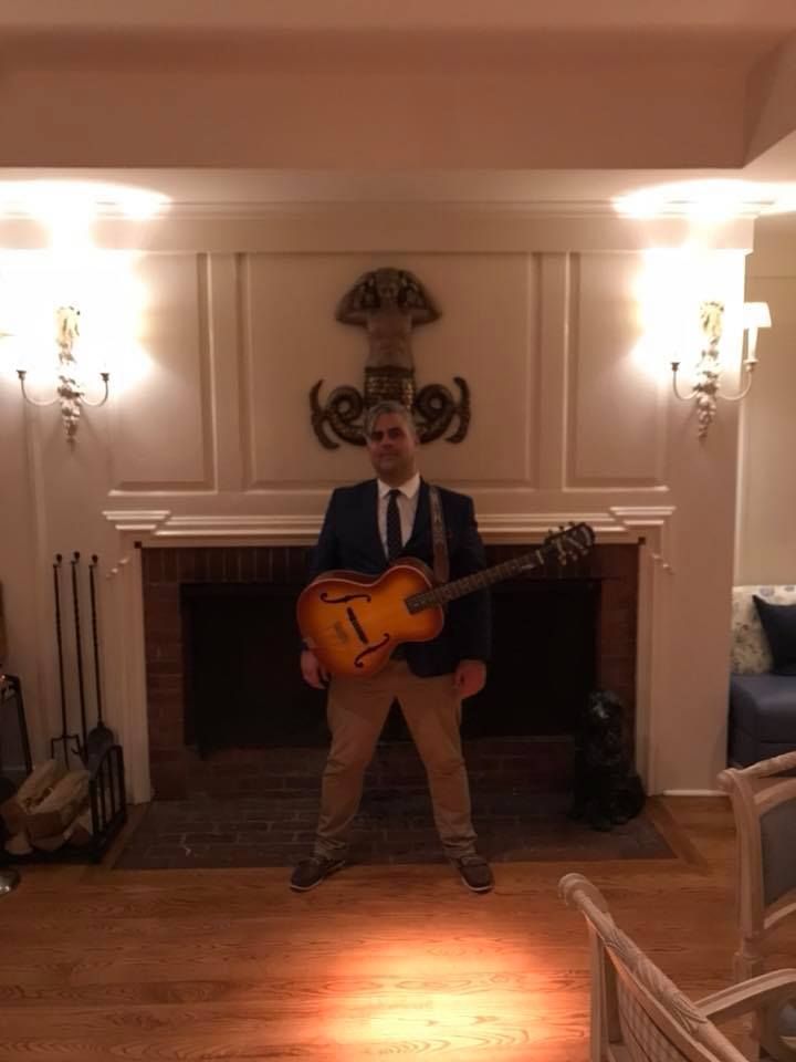 A man in a suit and tie is holding a guitar in front of a fireplace.